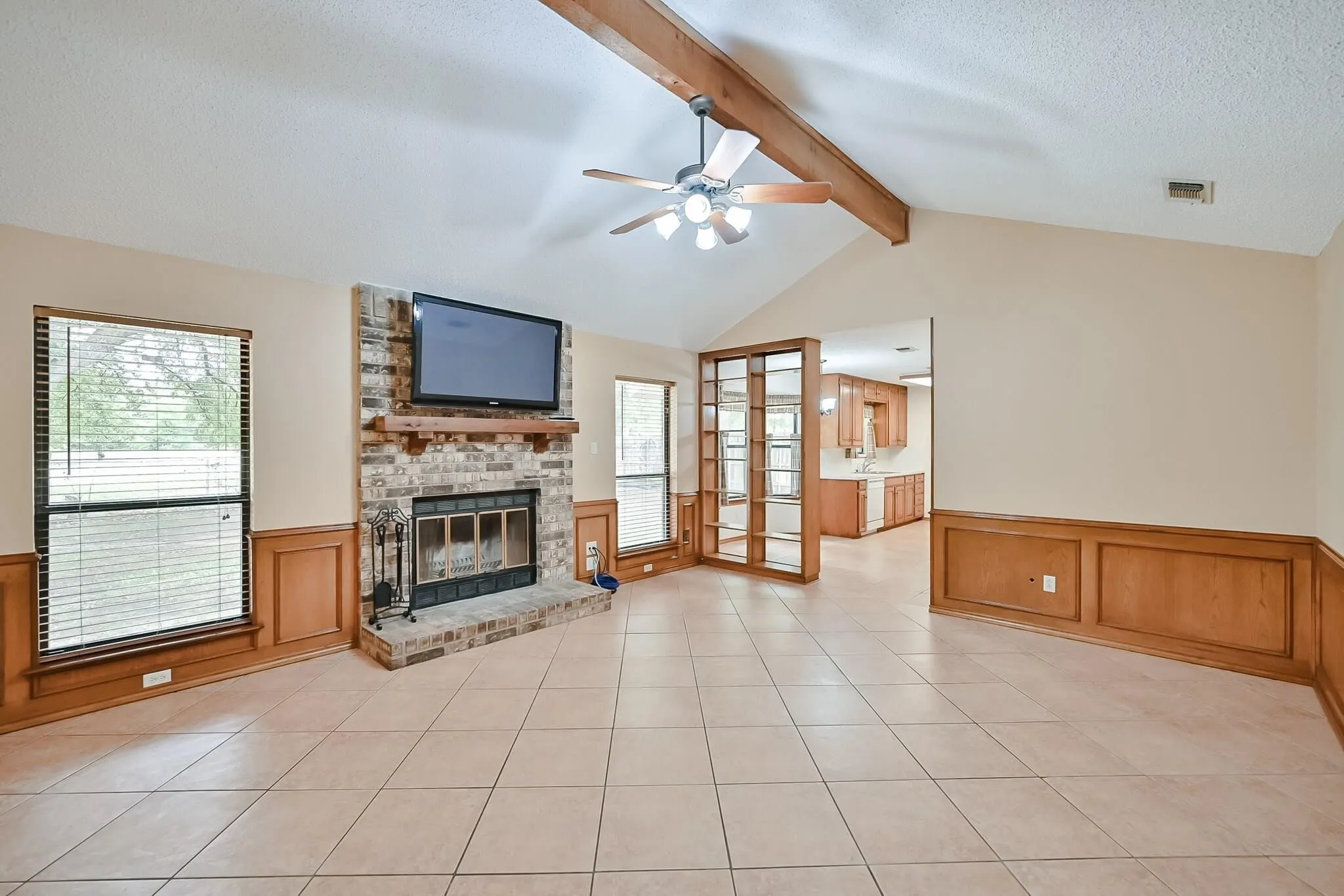 Vaulted ceiling with exposed wood beam, tiled flooring, wood paneling, brick fireplace with wood mantel, and a ceiling fan with light kit