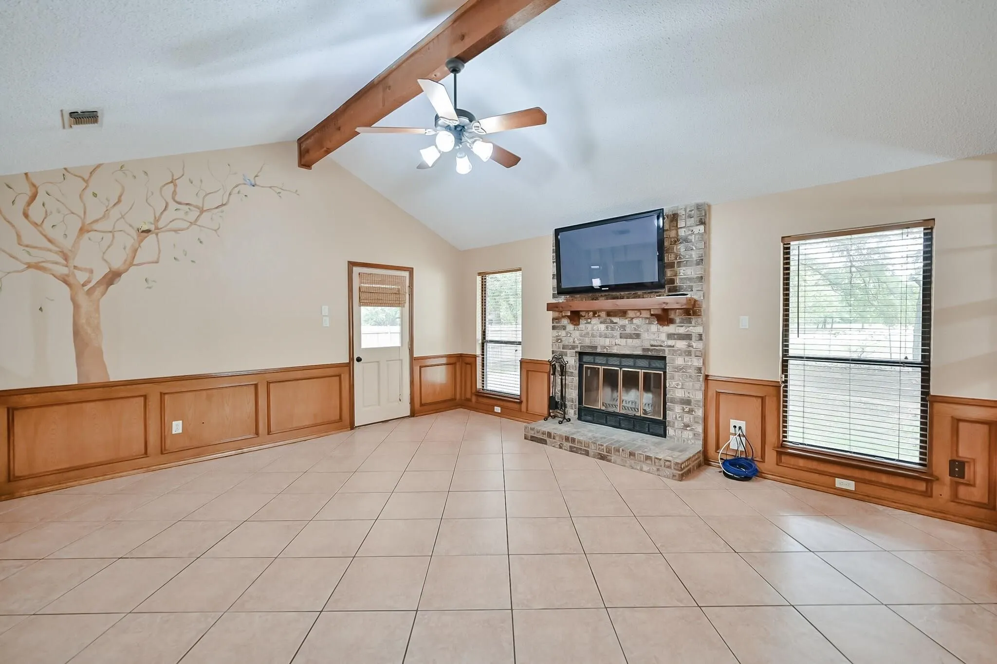 Vaulted ceiling with exposed wood beam and ceiling fan, corner brick fireplace with wood mantel, tile flooring, and wood paneling on lower walls