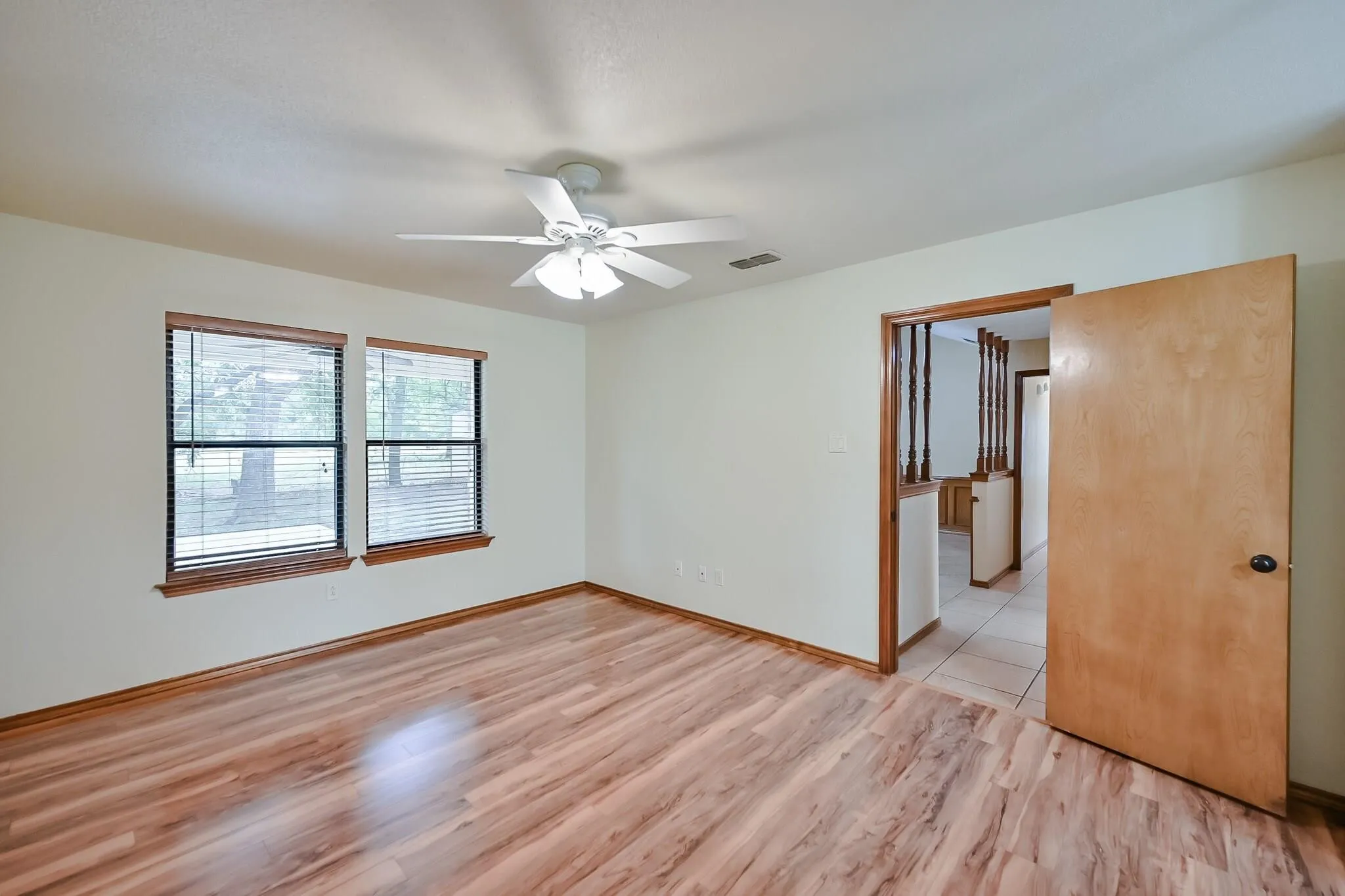 Spacious room featuring wood-finish flooring, two windows with blinds, a ceiling fan with integrated lighting, and a doorway with wood trim