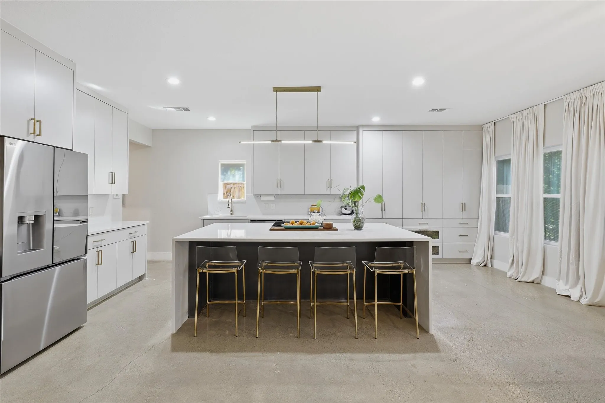 Sleek kitchen featuring an expansive central island with a waterfall edge, stainless steel refrigerator, abundant white cabinetry with gold-tone hardware, and recessed lighting