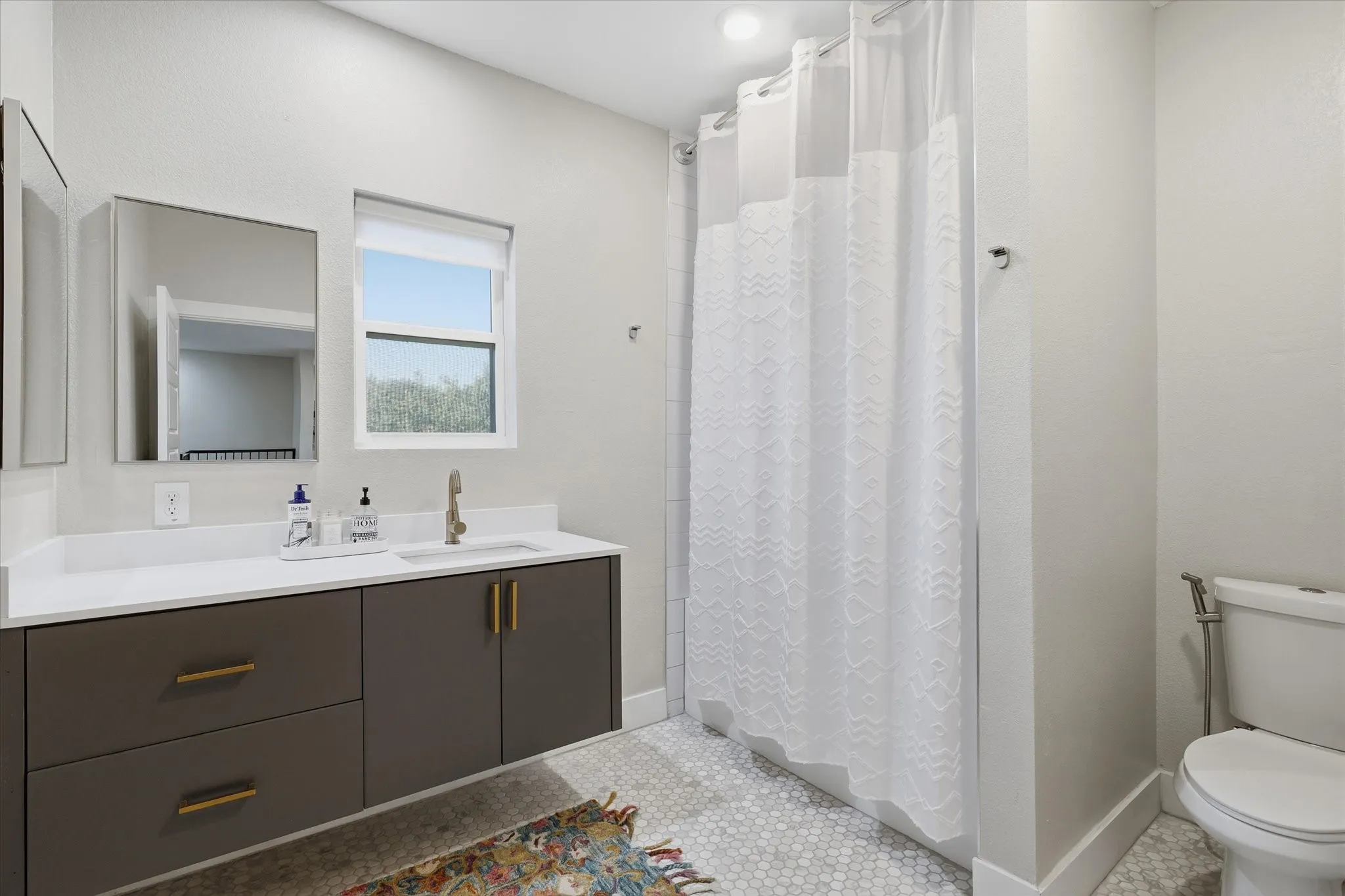 White hexagonal tile flooring, vanity with brass hardware, large framed mirror, and a window for natural light