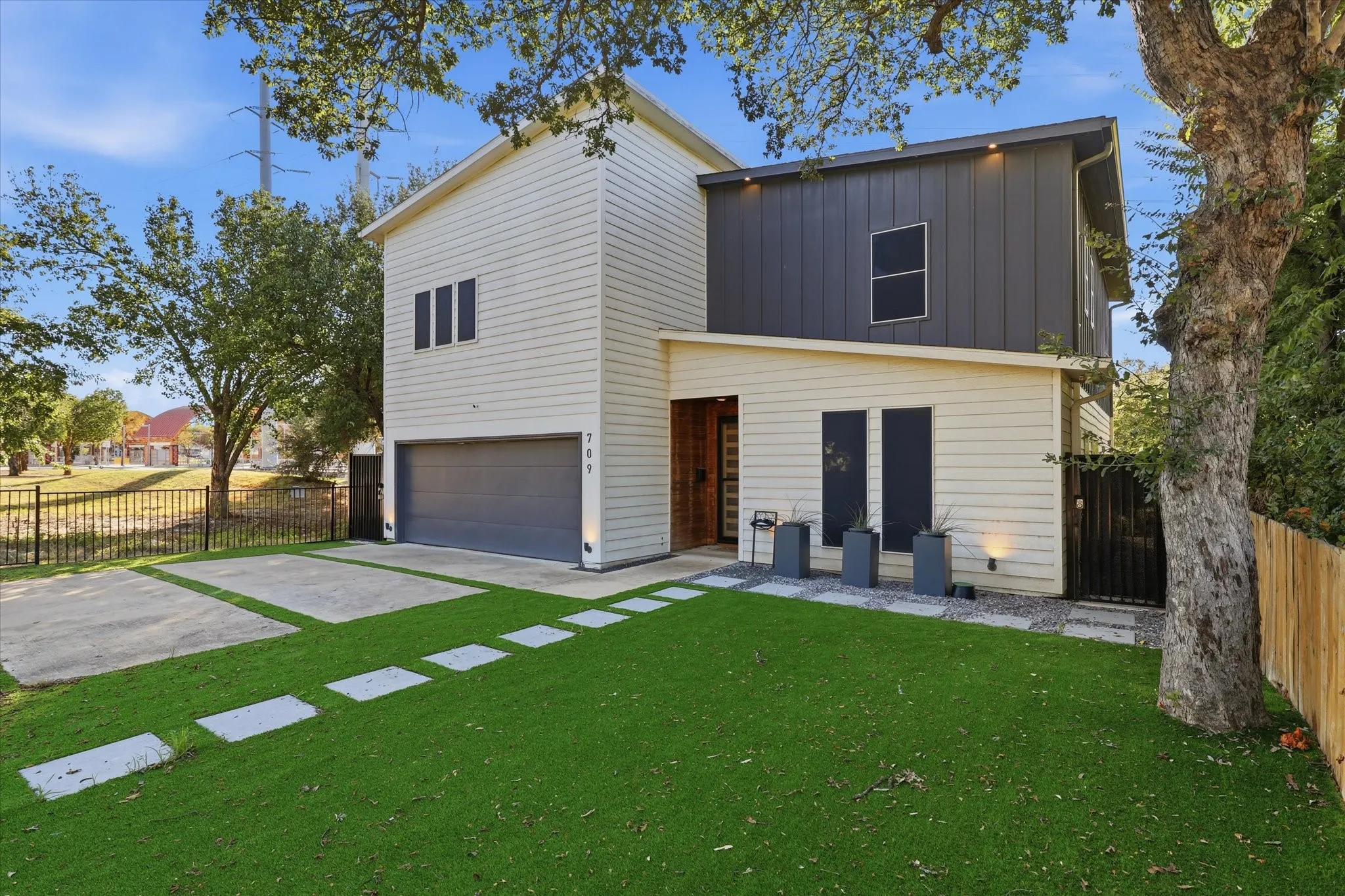 Modern exterior with light siding and vertical dark metal paneling, integrated garage, concrete driveway with green accents, and a fenced perimeter