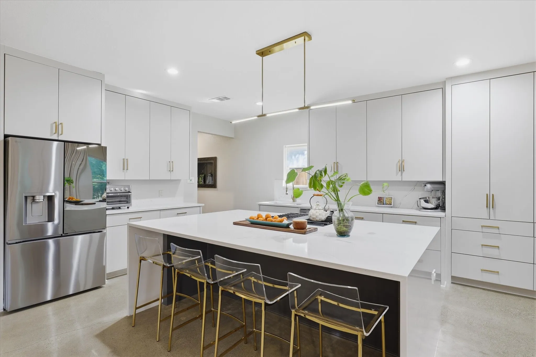 White cabinetry with gold-tone hardware, stainless steel refrigerator, white countertops, kitchen island with seating, and a linear pendant light fixture