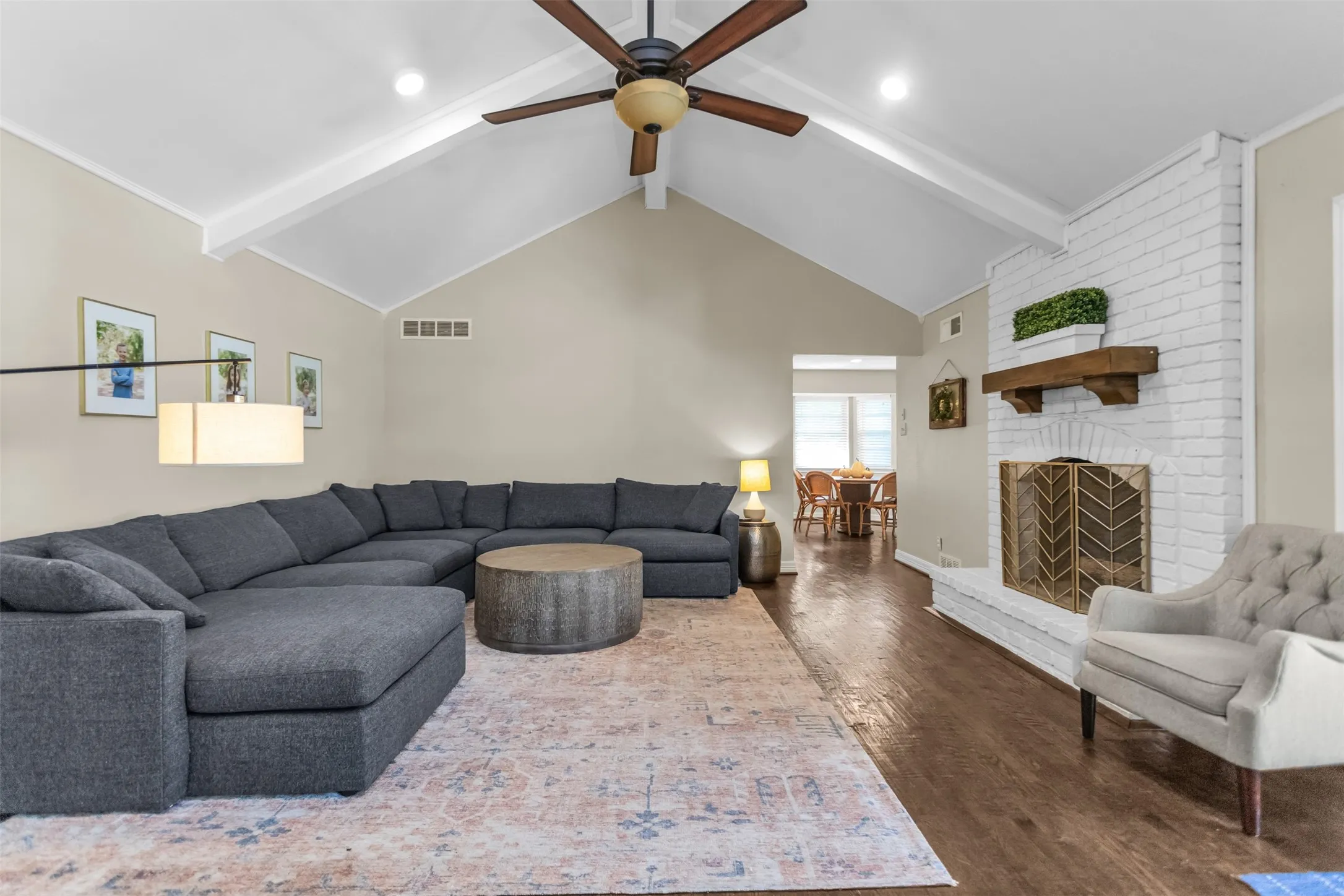 Vaulted ceiling with exposed beams, wood-finish flooring, white brick fireplace with a wood mantle, ceiling fan, and recessed lighting