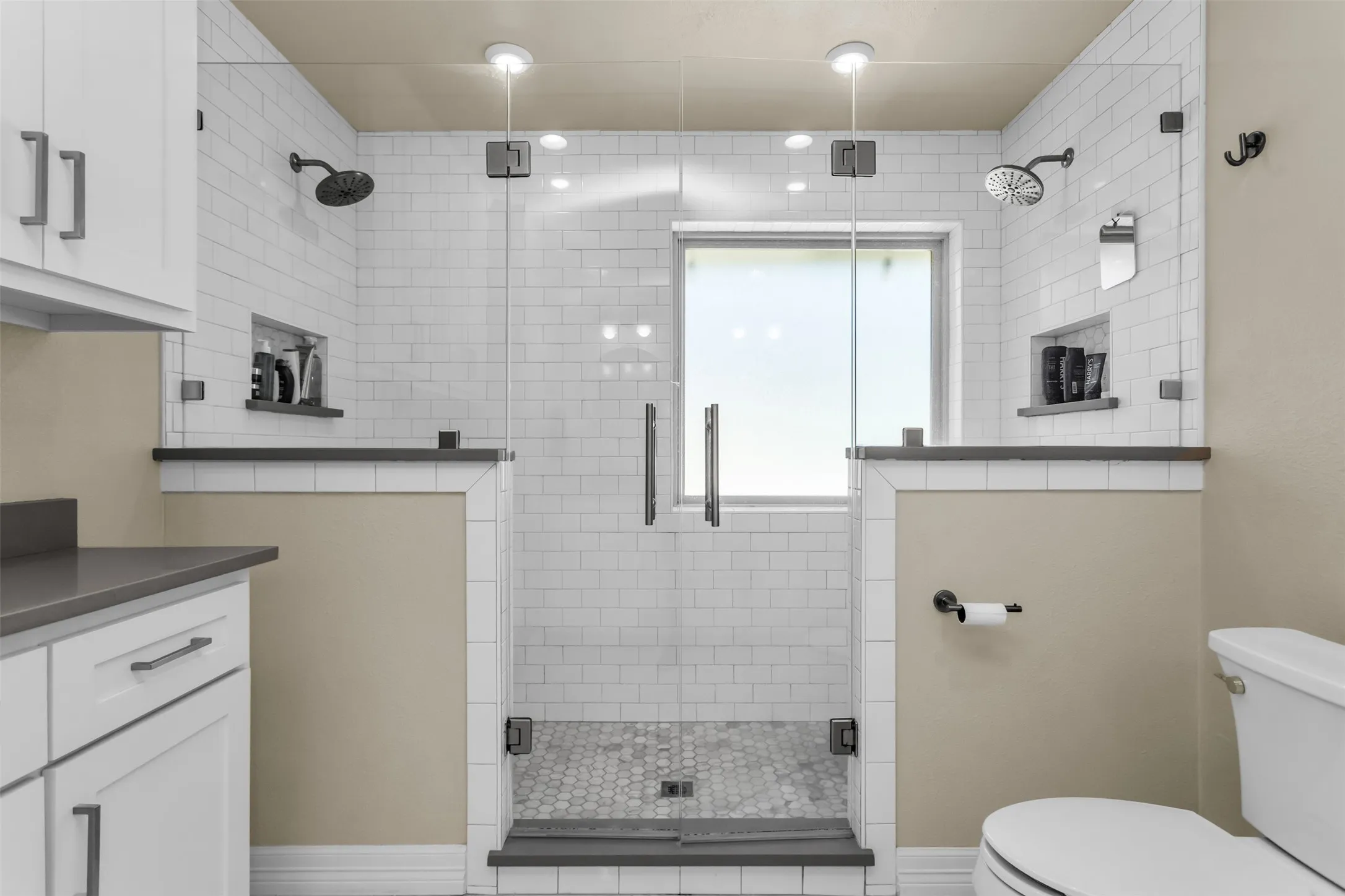Primary bathroom featuring a frameless glass-enclosed shower with dual showerheads, mosaic tile flooring, white subway tile walls, dual vanities with integrated countertops, and brushed nickel hardware throughout