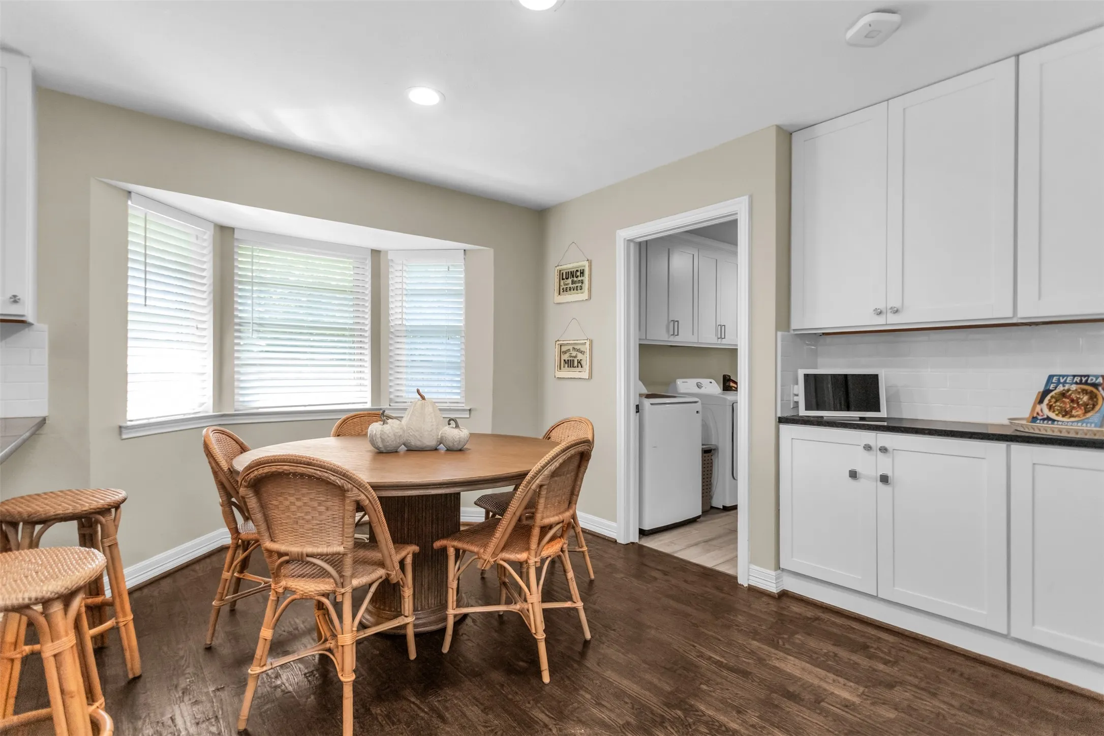 Wood-finish flooring, white shaker-style cabinetry, dark countertops, white subway tile backsplash, and a bay window with blinds