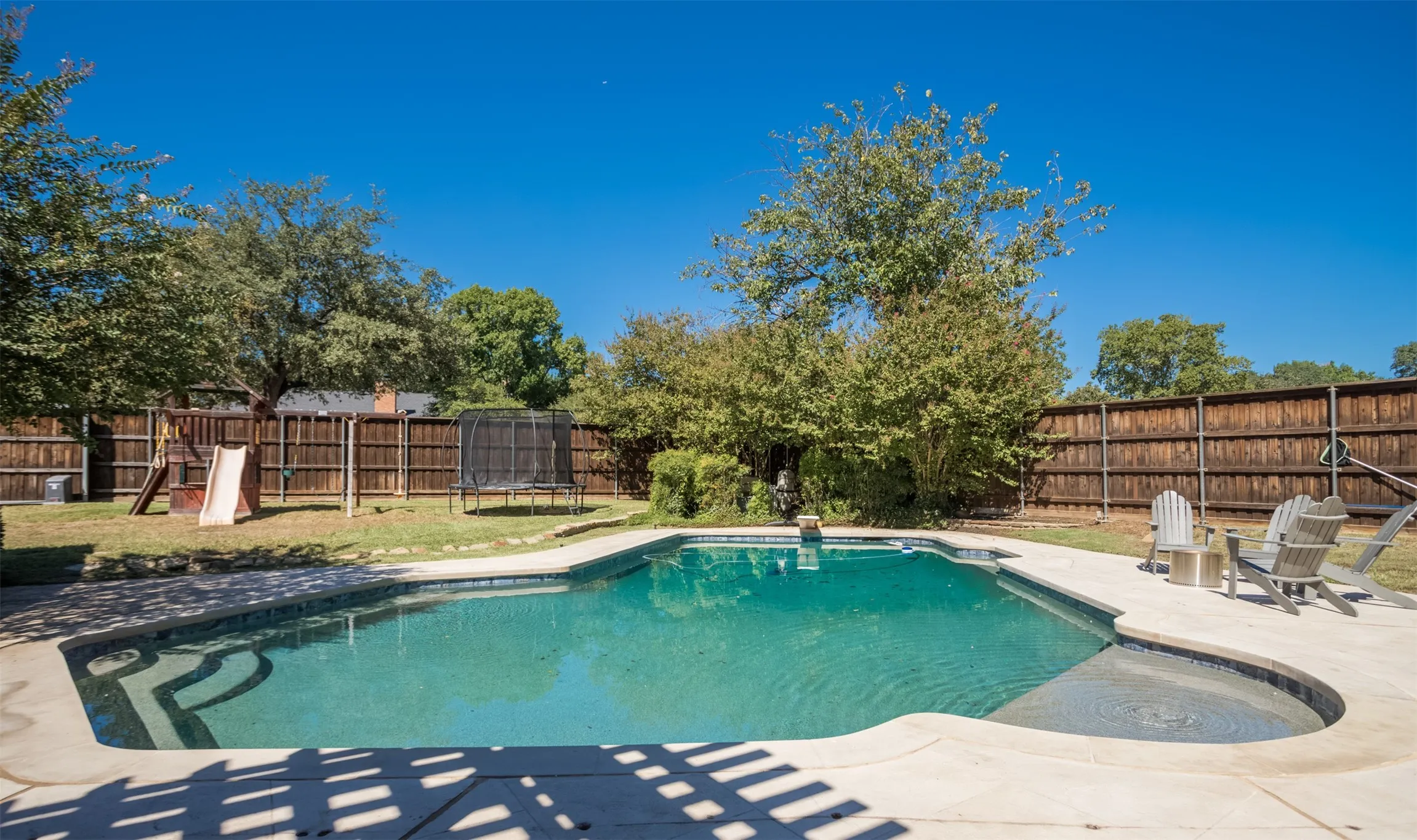 Resort-style in-ground swimming pool with sun shelf and spa, surrounded by mature landscaping and wood privacy fencing