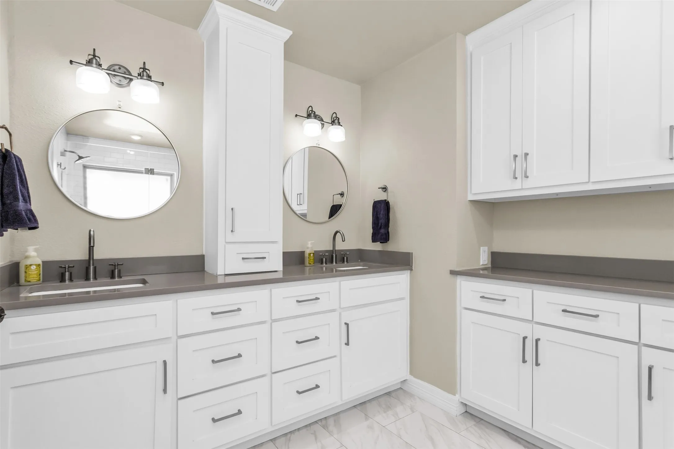 Dual-vanity primary bathroom featuring white shaker-style cabinetry, gray quartz countertops, polished round mirrors, chrome sconces, and tile flooring