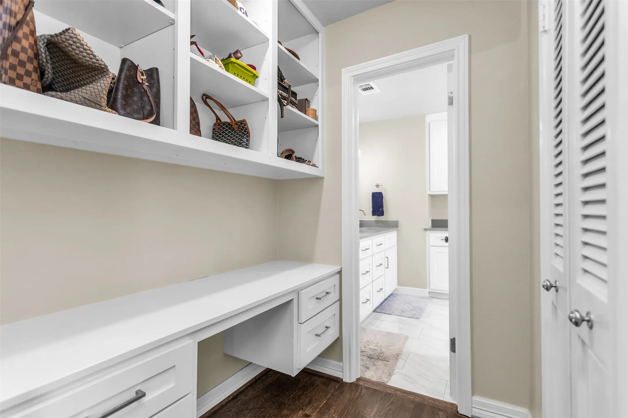 Elegant walk-in closet featuring custom white cabinetry with integrated drawers, expansive shelving with cubby storage for accessories, a dedicated work surface with ample counter space, wood-finish flooring, and direct access to the ensuite bathroom