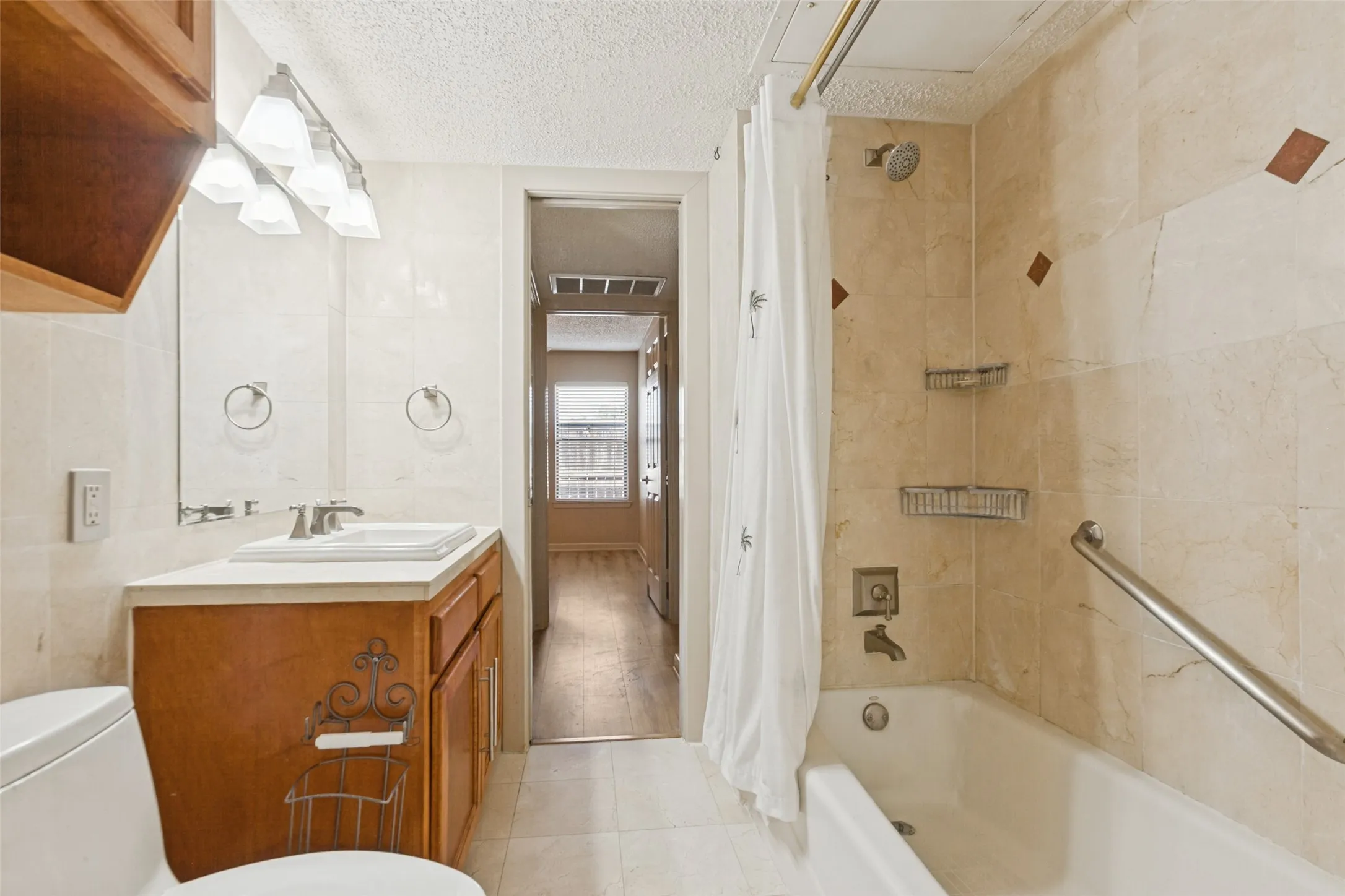 Tile flooring, wood-finish vanity with a built-in sink, wall-mounted mirror, and a tiled shower/tub combination with a grab bar