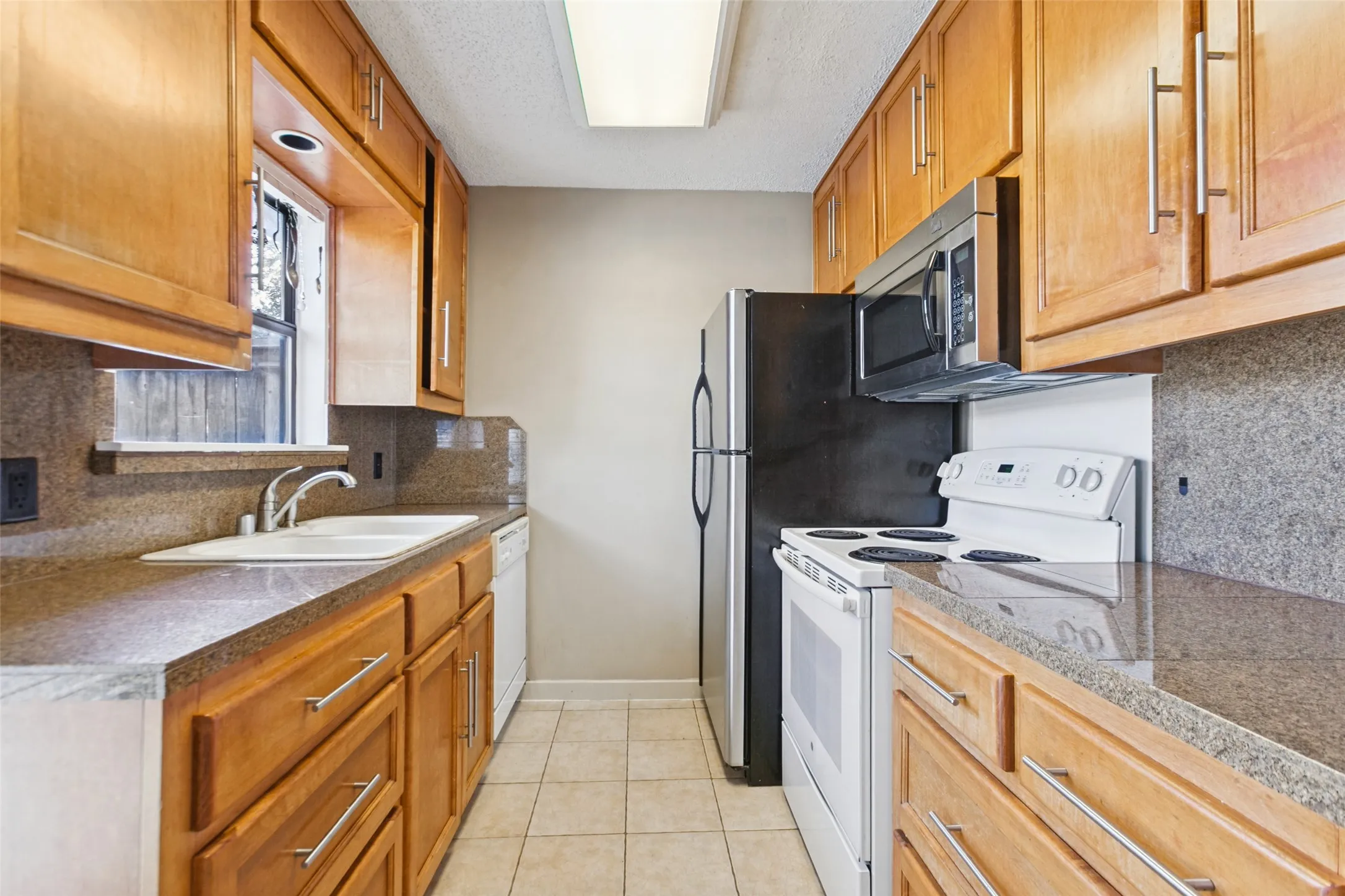 Galley kitchen featuring wood cabinetry, stone-finish countertops, tiled flooring, an undermount sink, and stainless steel appliances
