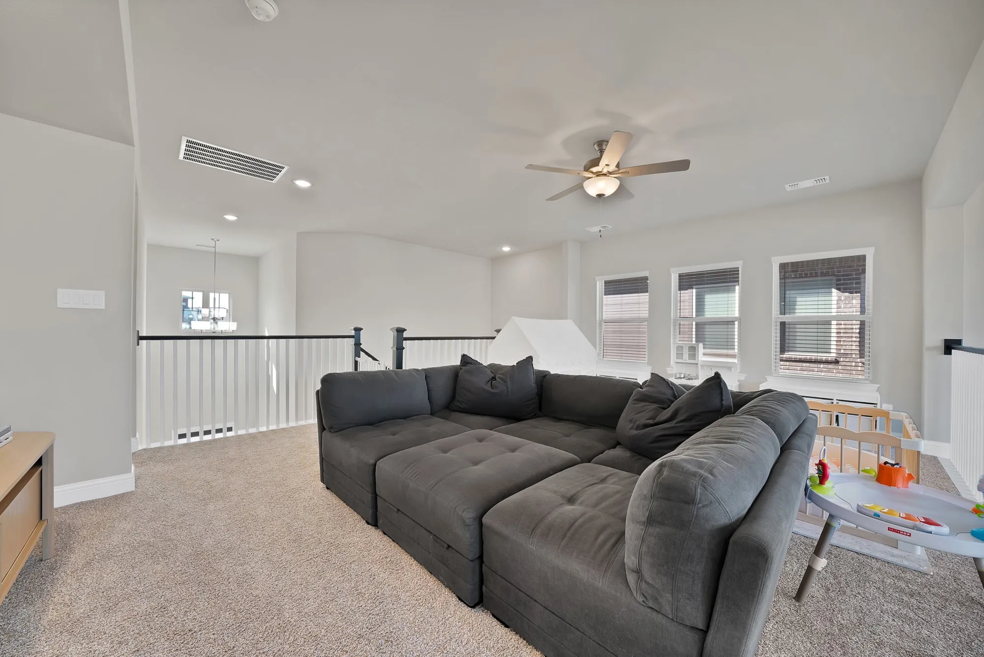 Carpeted loft space featuring a ceiling fan, three windows, and a white baluster railing with a black handrail