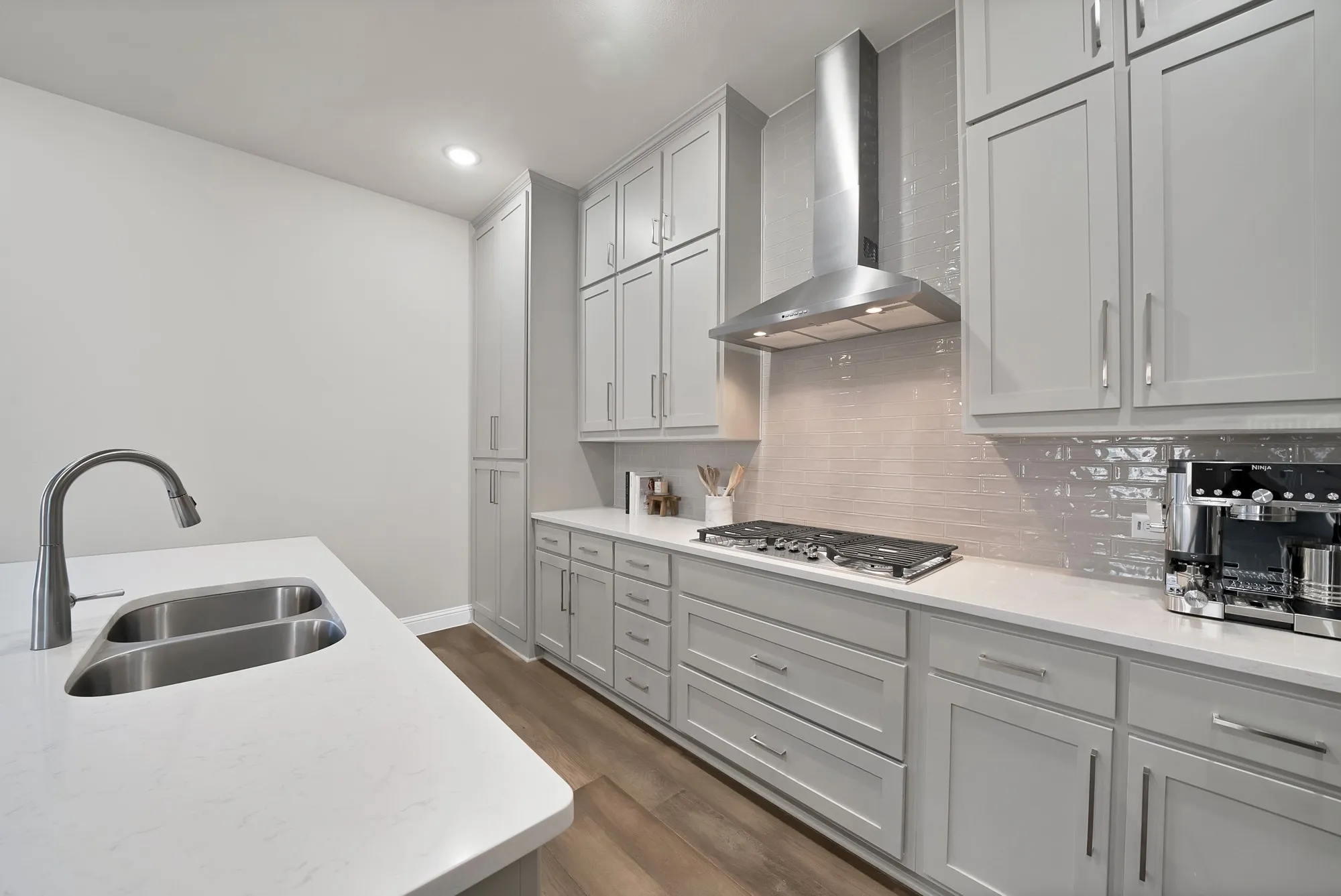 Gray shaker cabinetry, undermount double-basin sink, stainless steel range hood, stone-finish countertops, and wood-finish flooring
