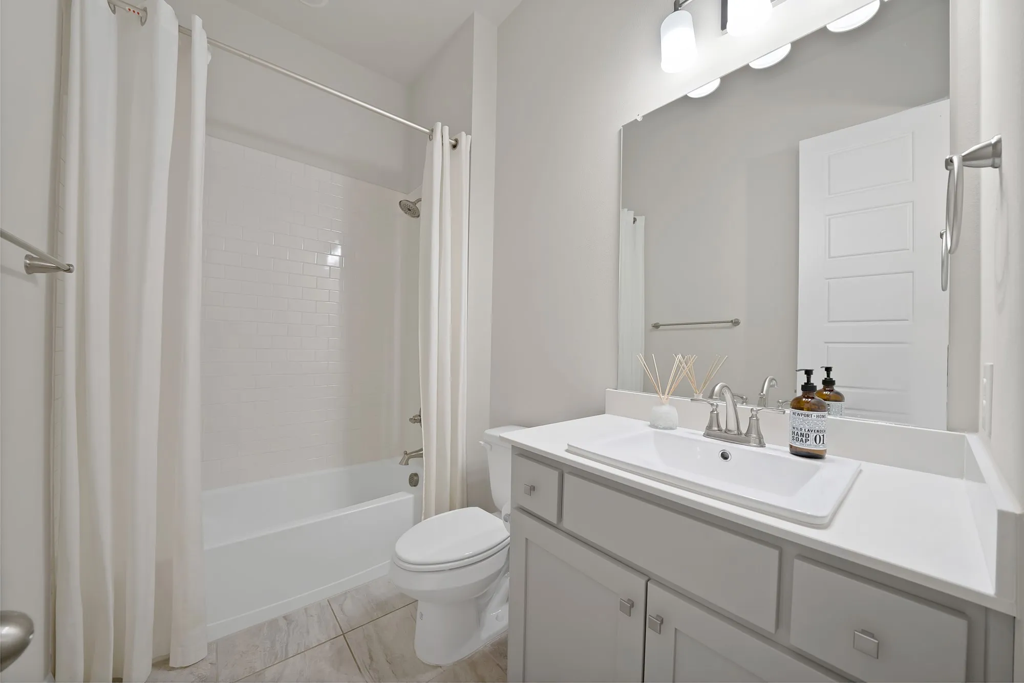 White subway tile bath surround, white tub, light gray vanity with rectangular integrated sink, neutral tile flooring, and brushed nickel hardware
