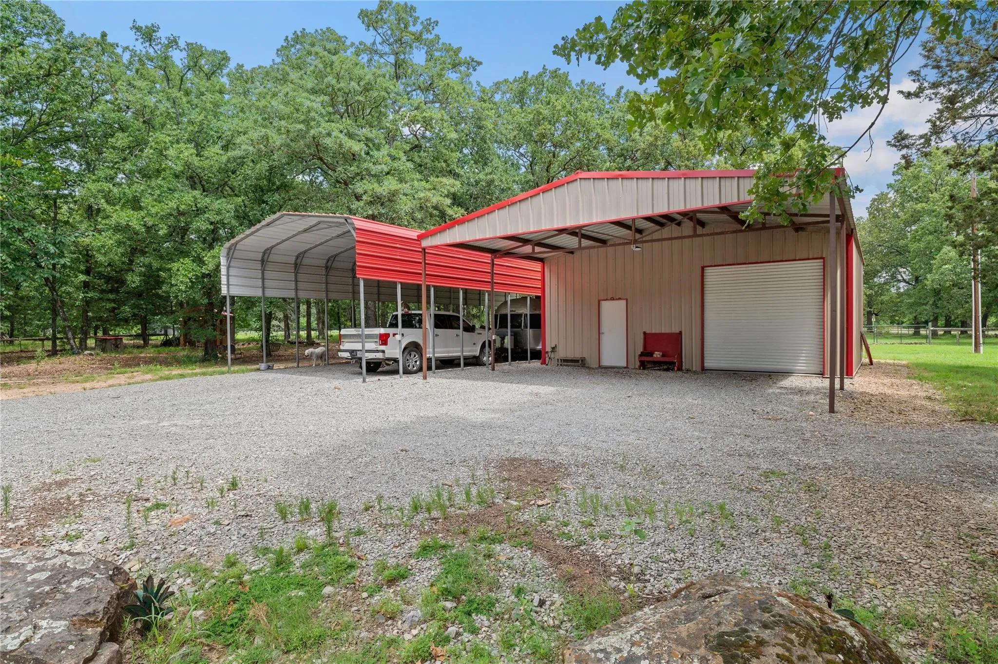 Metal-sided building featuring a red roof, a roll-up garage door, an entry door, and an attached RV carport with a gravel parking area.