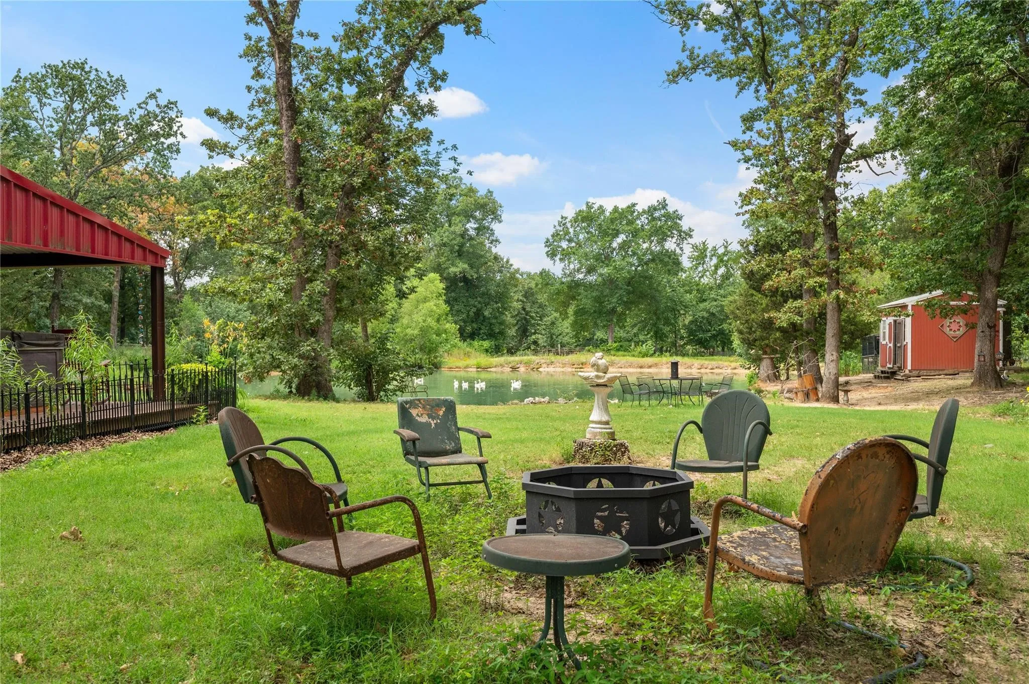 Expansive lawn area featuring a fire pit, mature trees, and visible pond with chicken coop in the background.