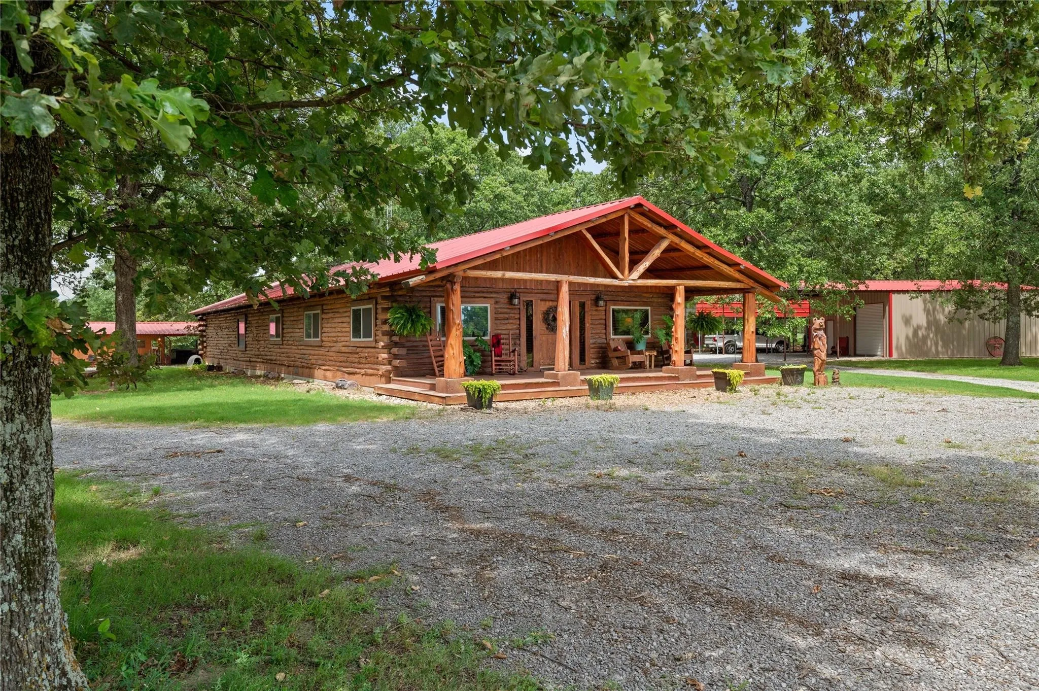 Rustic log cabin with a distinctive red metal roof and exposed beam detailing. Garage/Shop/RV parking in the back right. Outdoor living center in the back left.