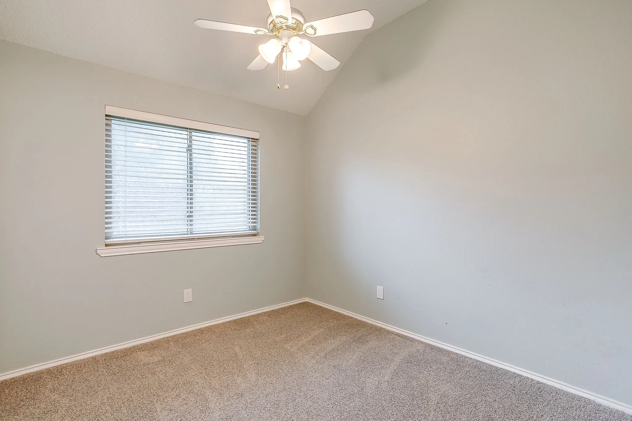 Vaulted ceiling, ceiling fan with light fixture, window with blinds, light-colored walls, and carpet flooring