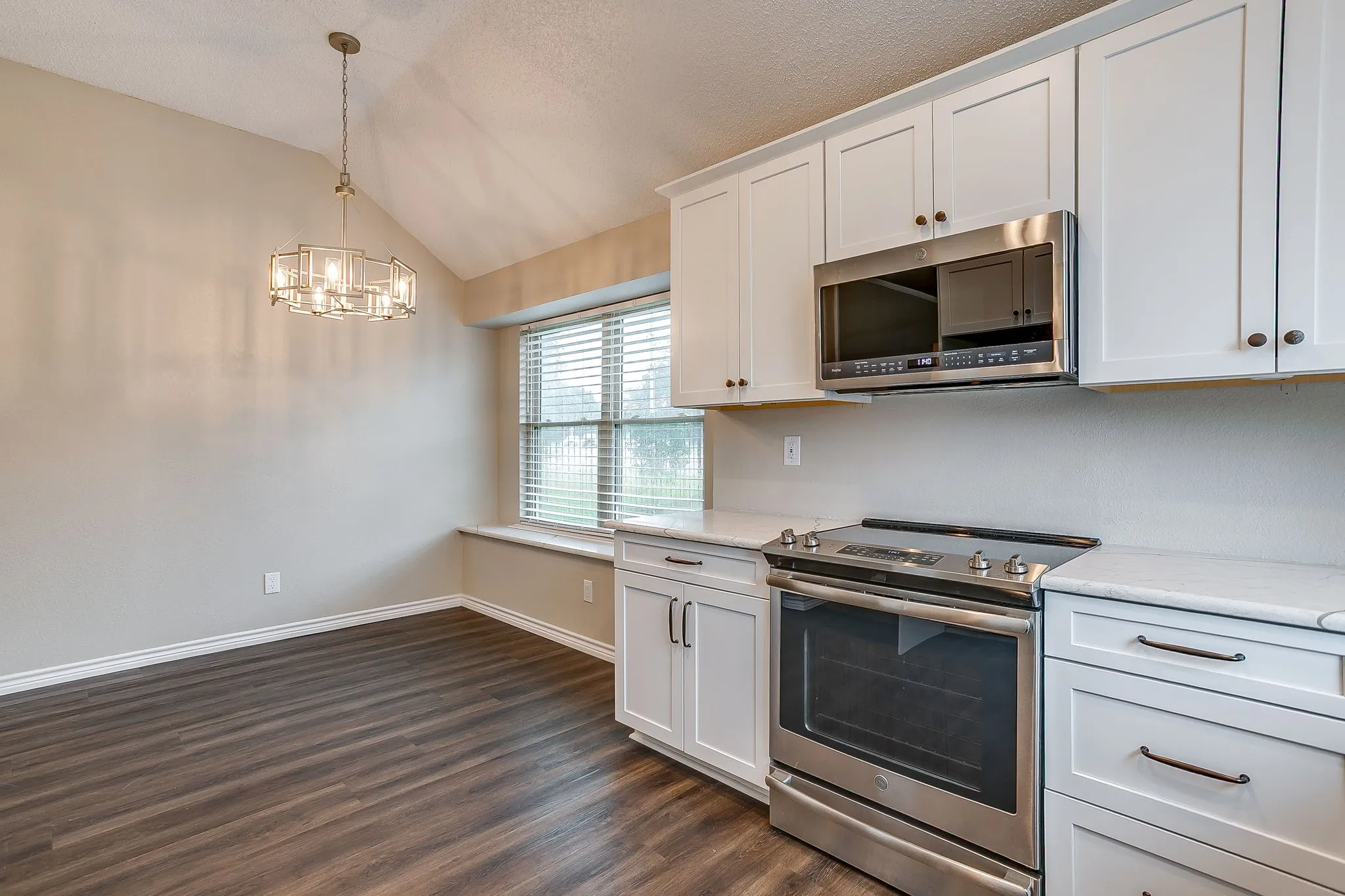 Vaulted ceiling with a geometric chandelier, dark wood-finish flooring, white cabinetry, stainless steel appliances, and light-toned countertops