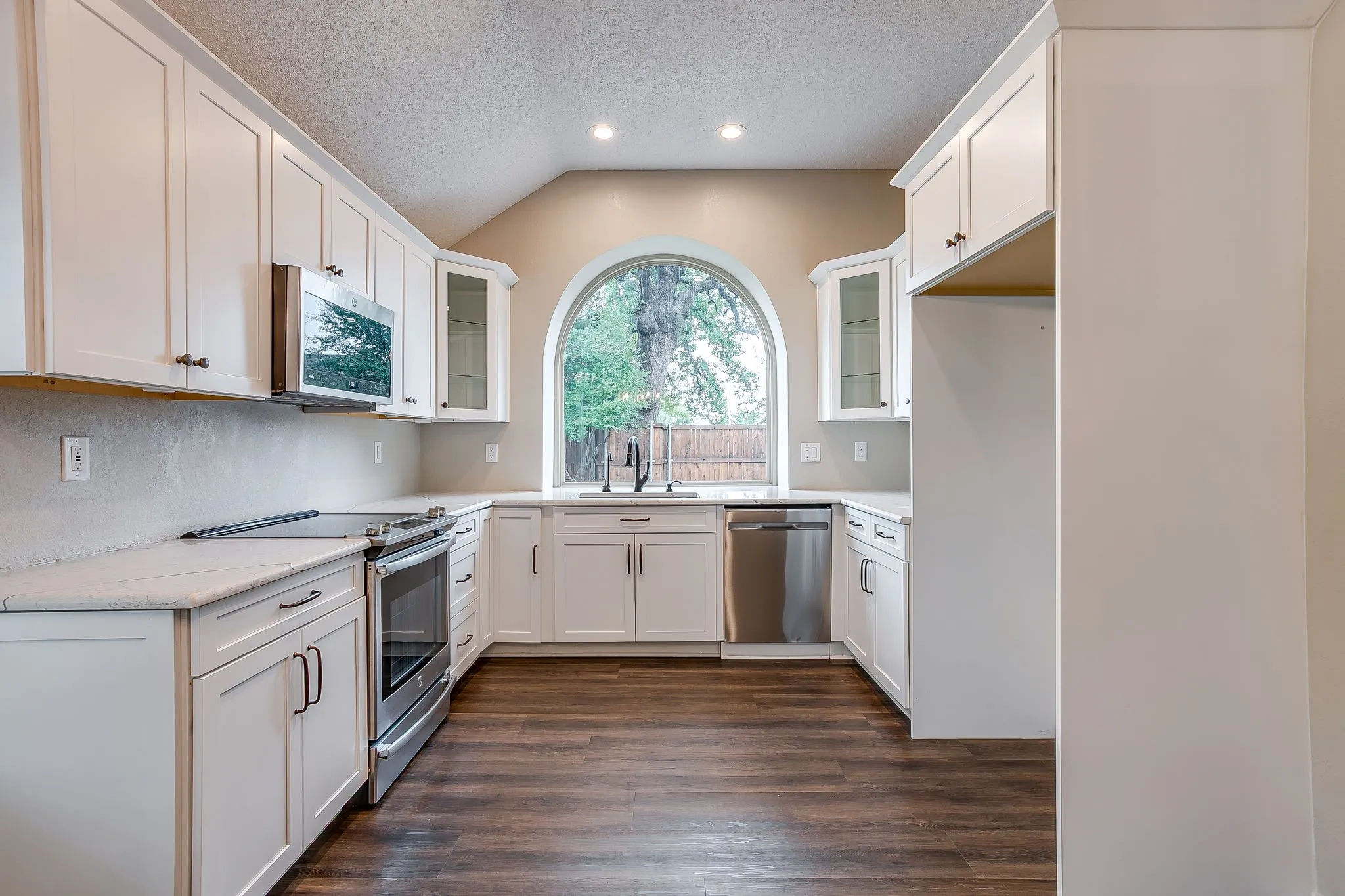 Kitchen featuring white shaker-style cabinetry, stainless steel appliances, light-toned countertops, wood-finish flooring, and an arched window