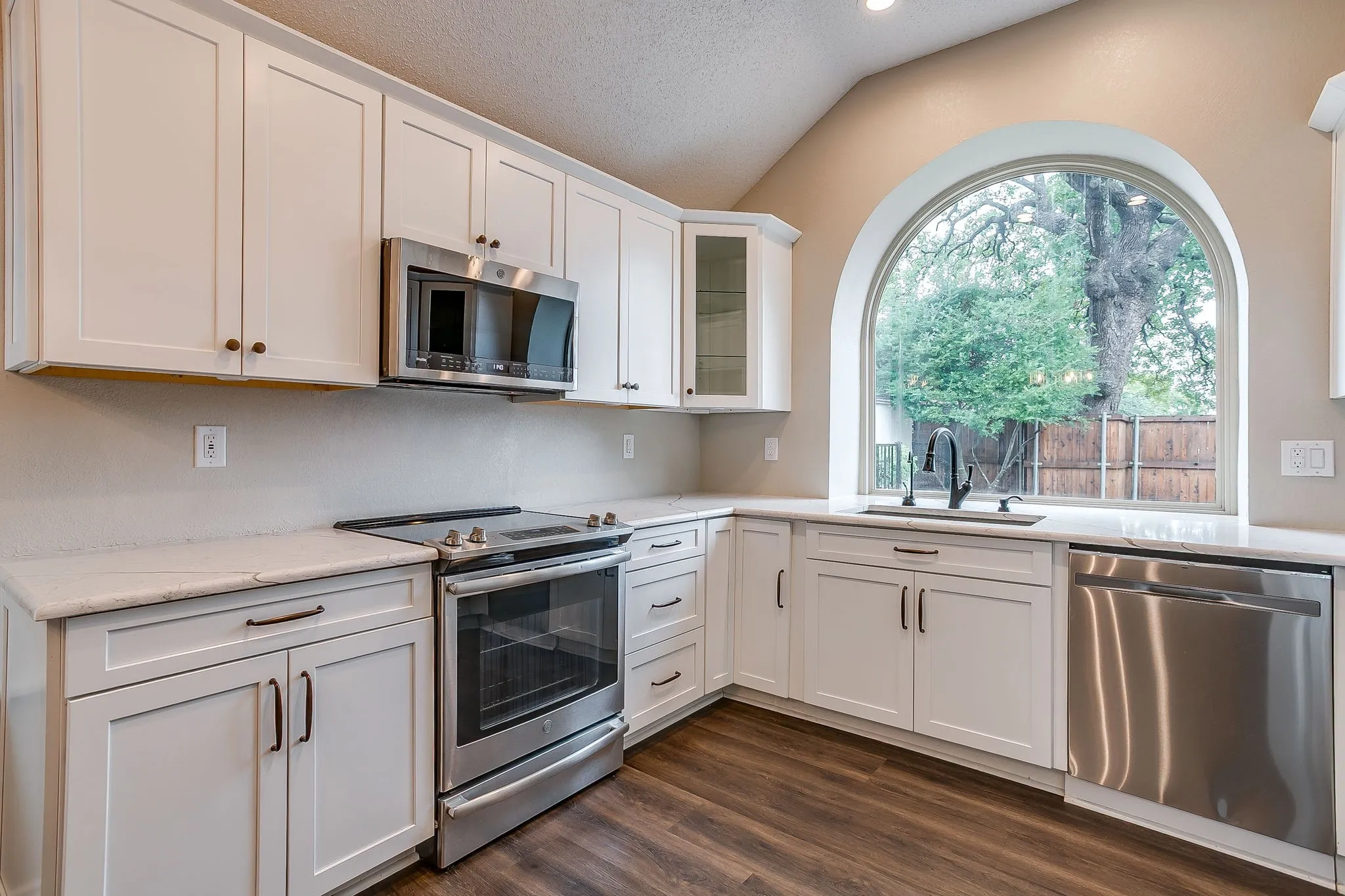 White shaker-style cabinetry with dark hardware, stainless steel appliances, stone countertops, wood-finish flooring, and an arched window