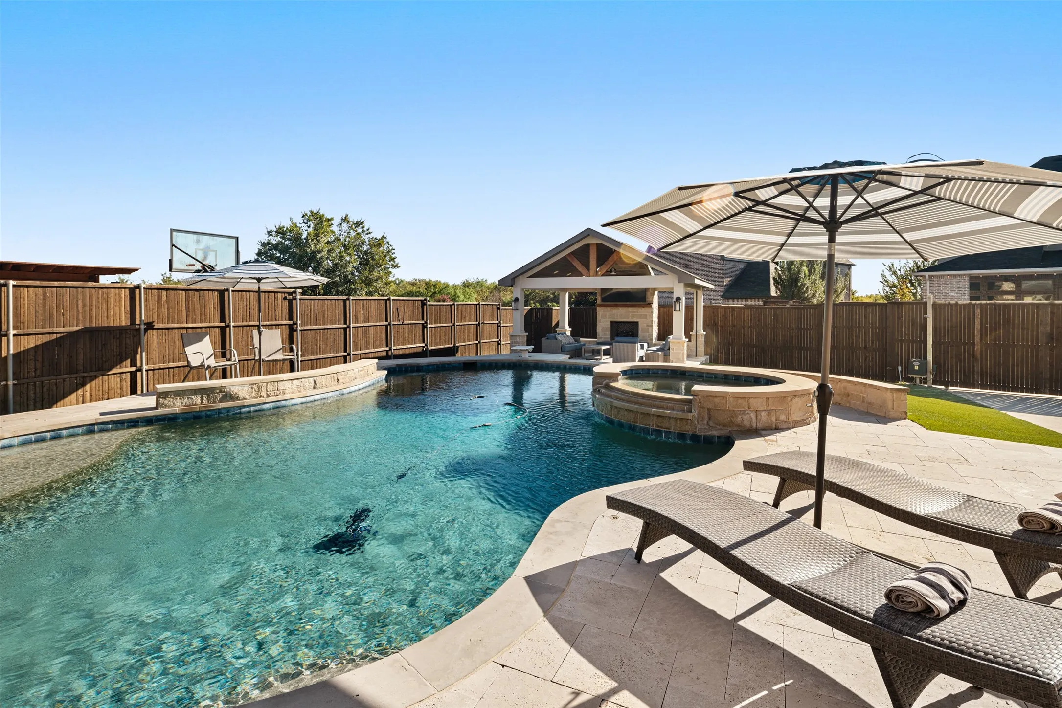 Curved swimming pool with integrated spa and stone surround, accompanied by an outdoor living structure featuring a fireplace and recessed lighting