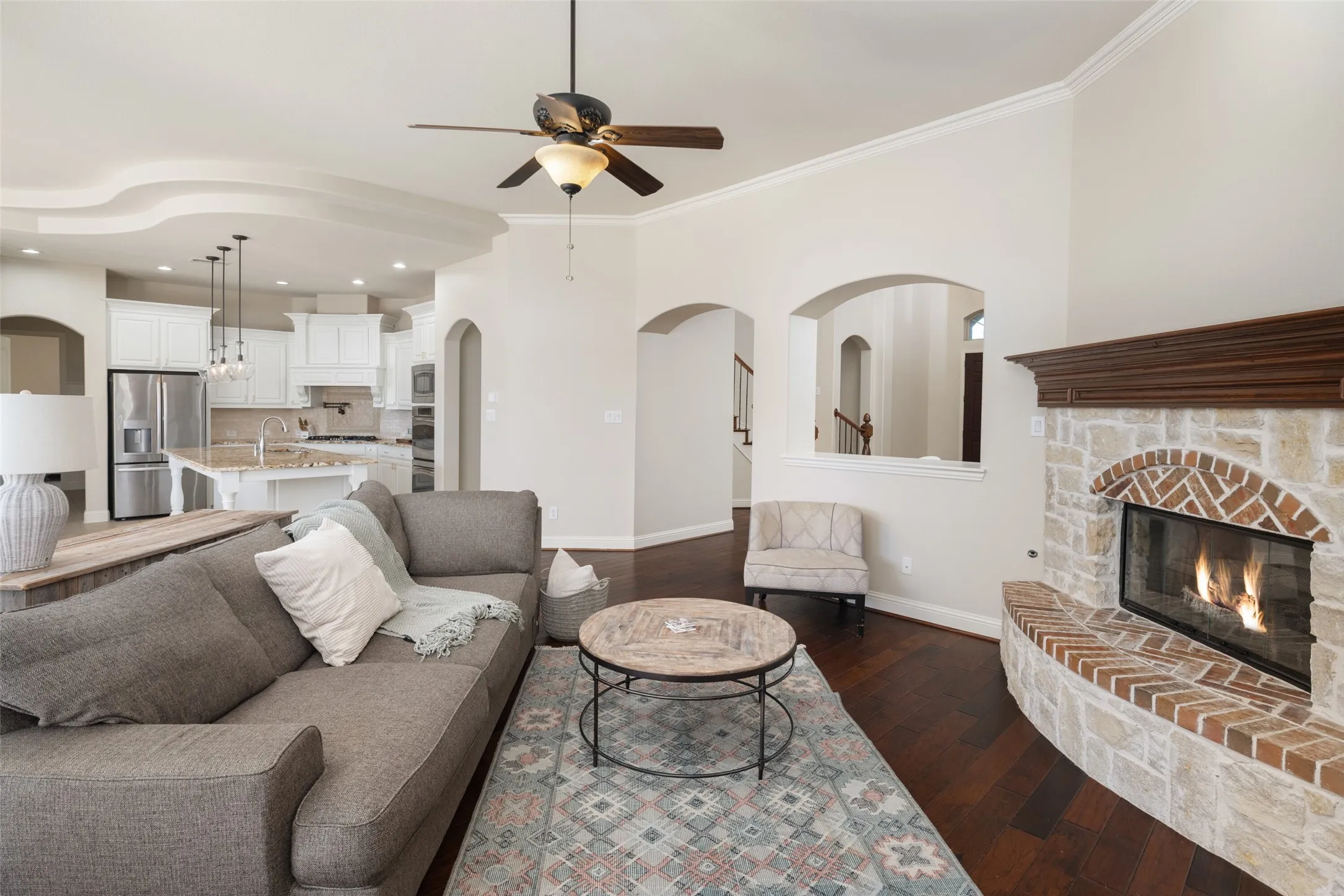 Living area featuring dark wood-finish flooring, a stone and brick fireplace with a wood mantle, and an open layout to a kitchen with white cabinetry and stainless steel appliances
