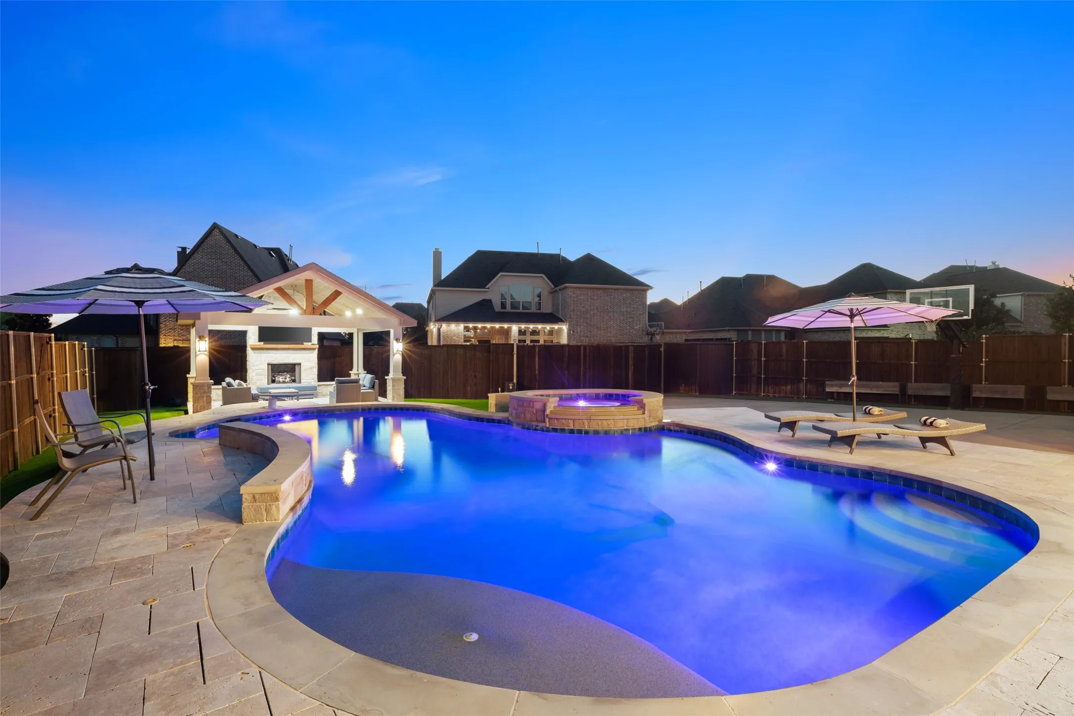 Expansive backyard featuring a custom-shaped pool with integrated spa, a built-in outdoor kitchen with fireplace, and paver stone hardscaping