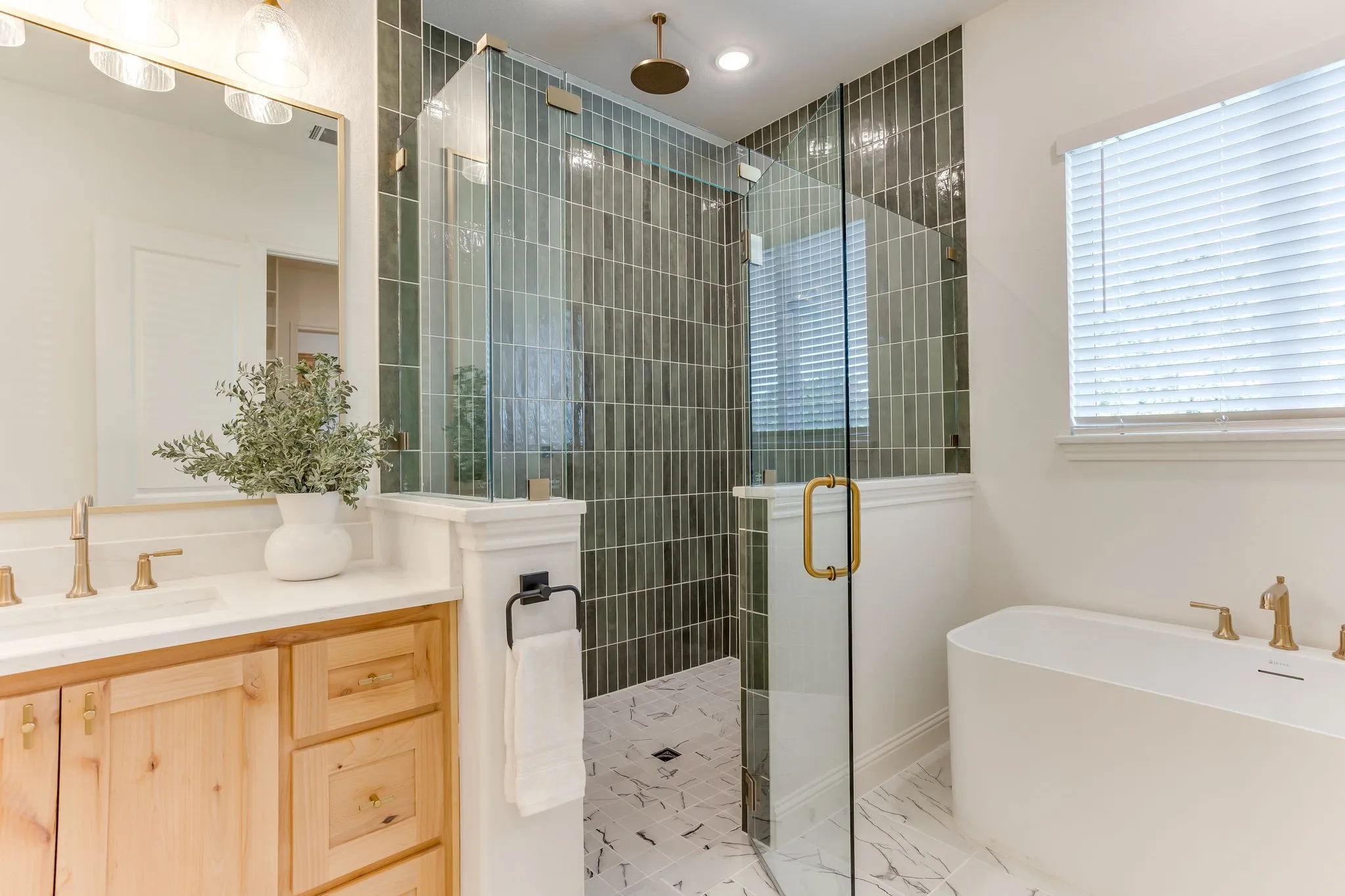 Walk-in shower with vertical tile detailing, freestanding soaking tub, wood-finish vanity with countertop, and gold-toned fixtures
