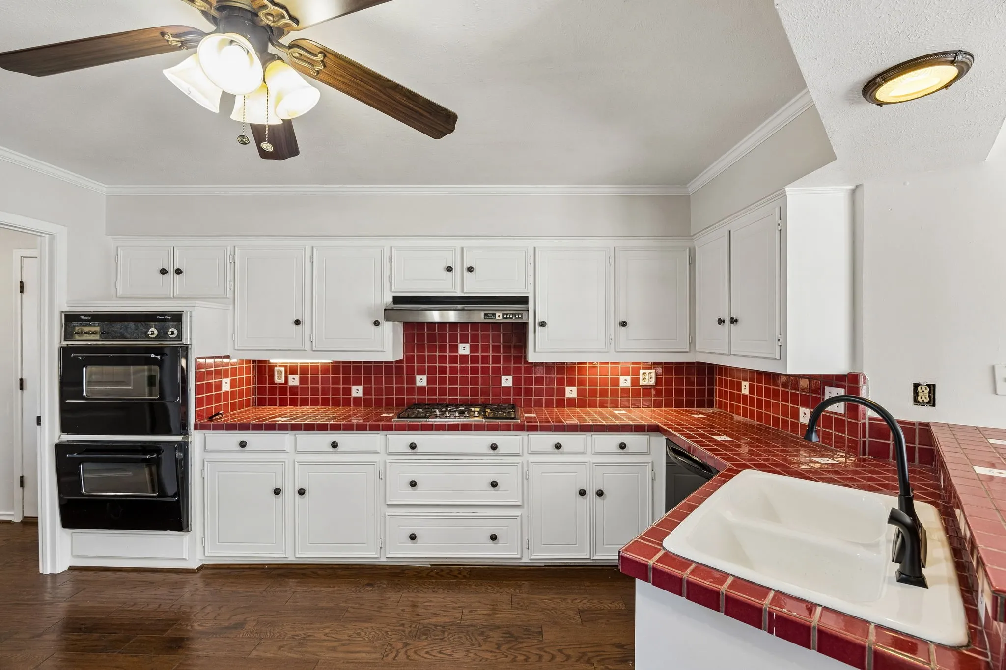 Kitchen featuring white cabinetry, red tile countertops, red tile backsplash, double wall ovens, with laundry room off to the side