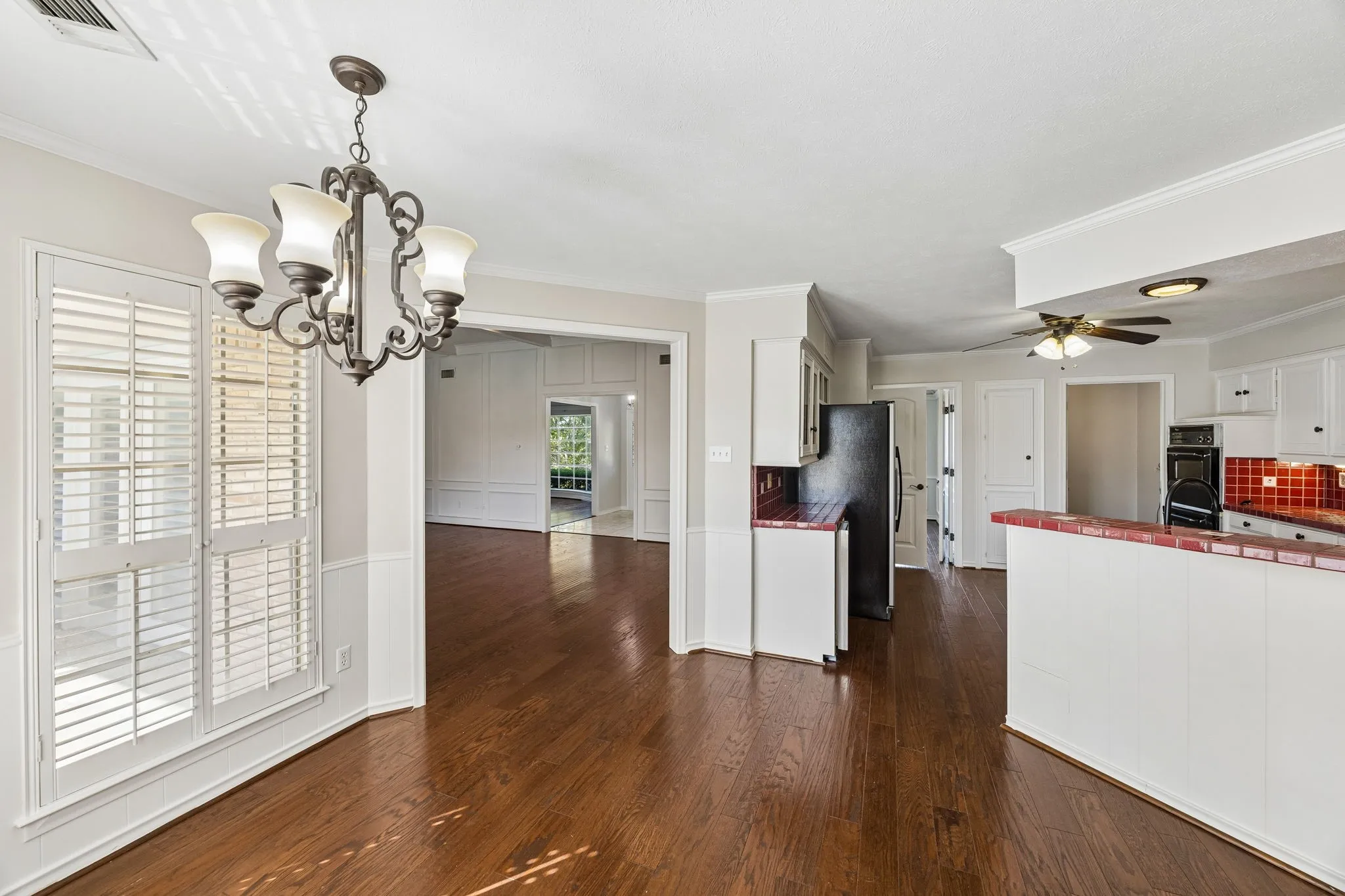 Wood-finish flooring throughout, with a decorative chandelier, white plantation shutters, and a kitchen featuring red tile accents