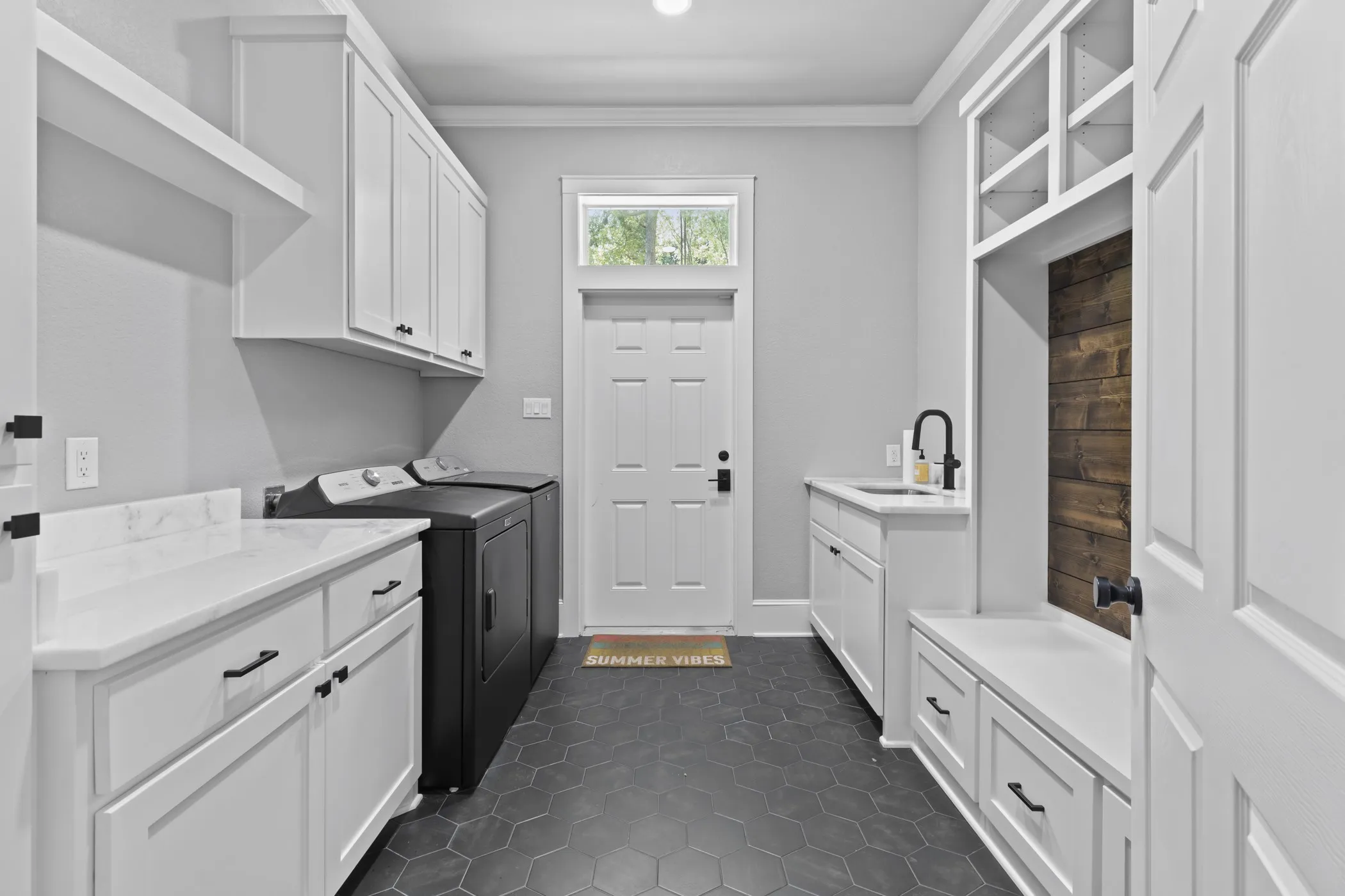 White shaker cabinetry with black hardware, white countertops, built-in mudroom bench with storage, black hexagonal floor tile, and a single basin sink with a black faucet