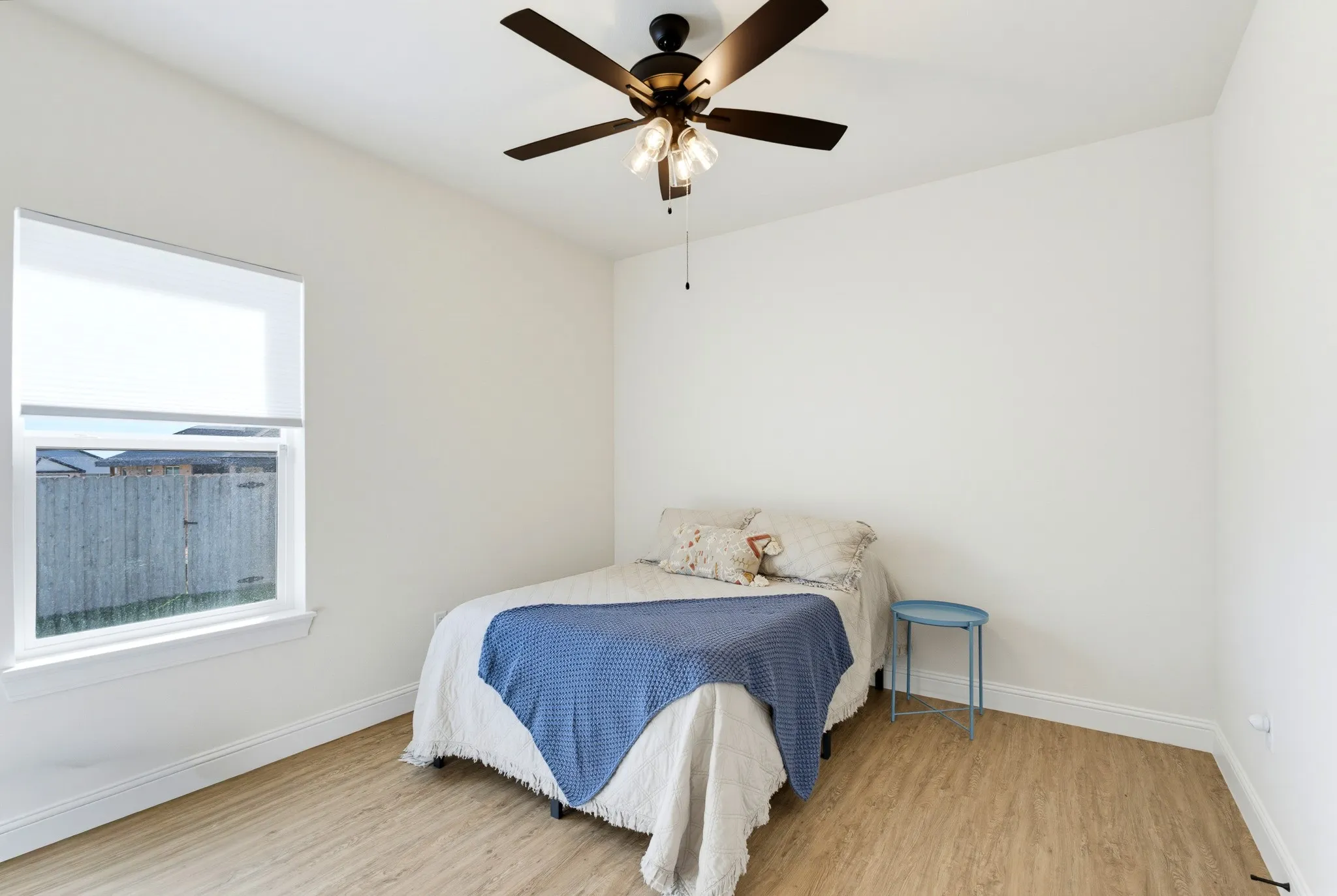 Bright room with wood-finish flooring, a ceiling fan with integrated lighting, and a window featuring a roller shade