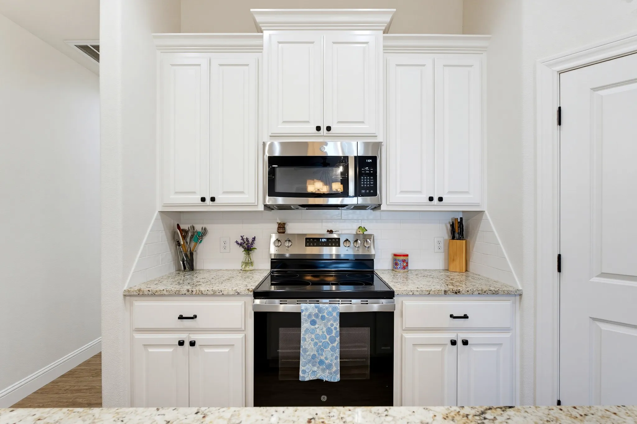 White shaker-style cabinetry with black hardware, stainless steel range with overhead microwave, and light-toned speckled countertops with white subway tile backsplash