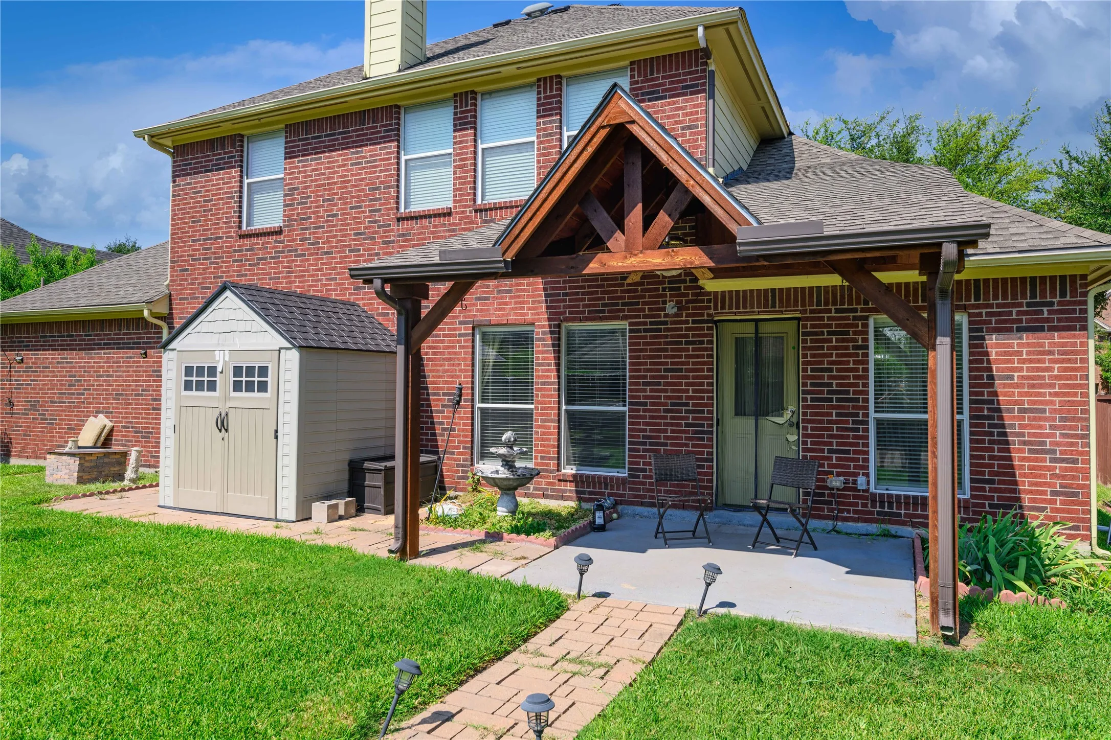 Brick exterior featuring a gabled roof, covered patio with wood beam accents, grass lawn, and an exterior storage shed