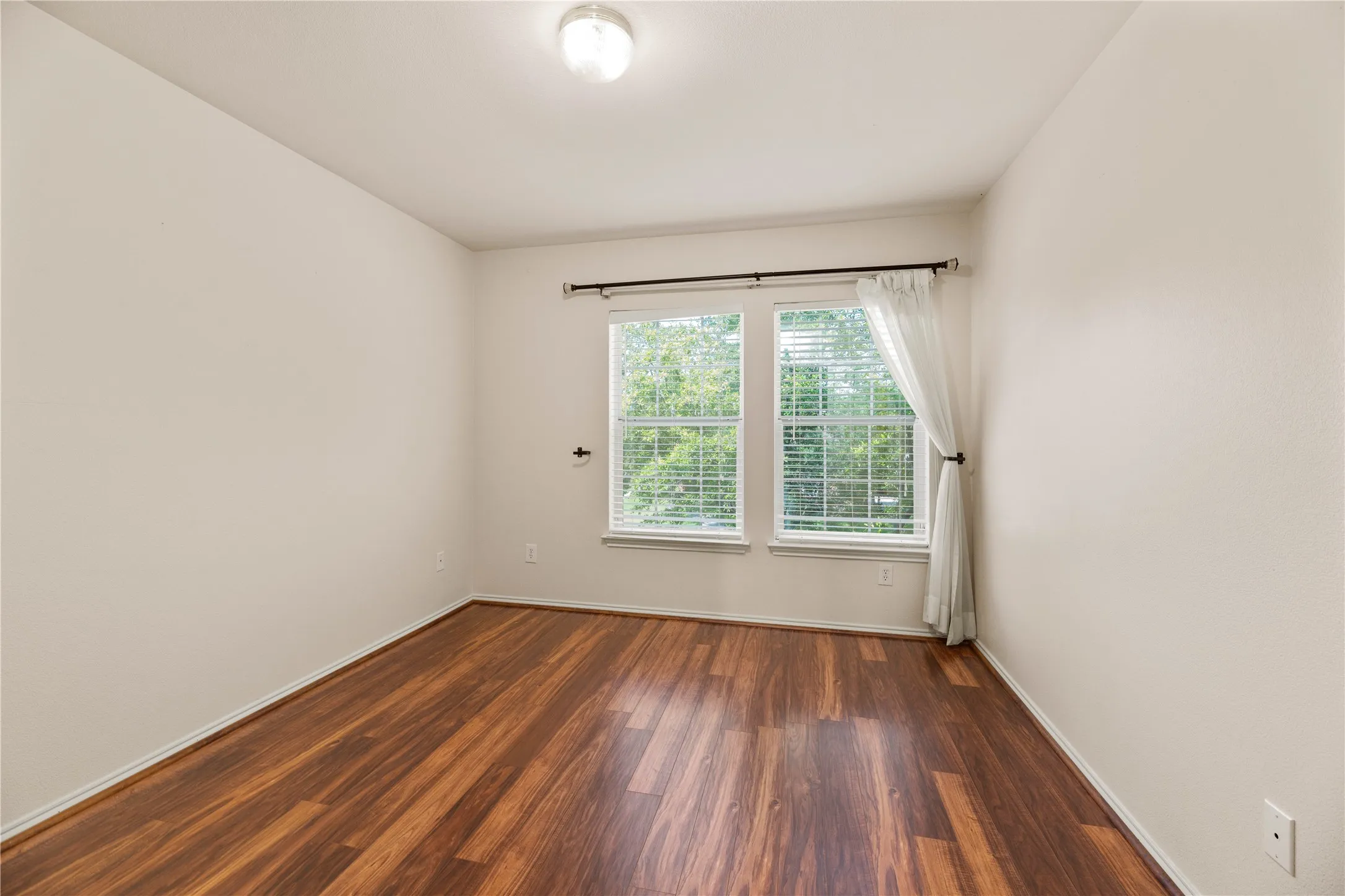 Wood-finish flooring with a satin sheen, dual windows with blinds, ceiling-mounted light fixture, and neutral wall paint