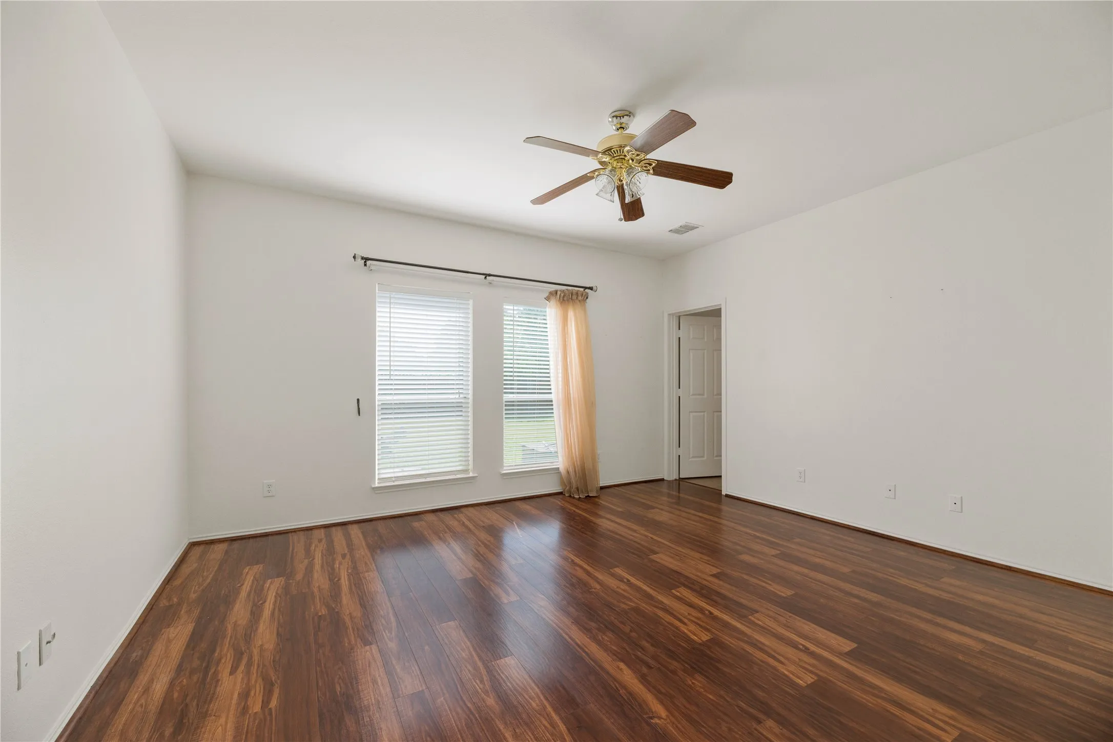 Spacious room flooded with natural light through dual glass doors and windows, featuring rich wood-finish flooring throughout and a ceiling fan with brass accents