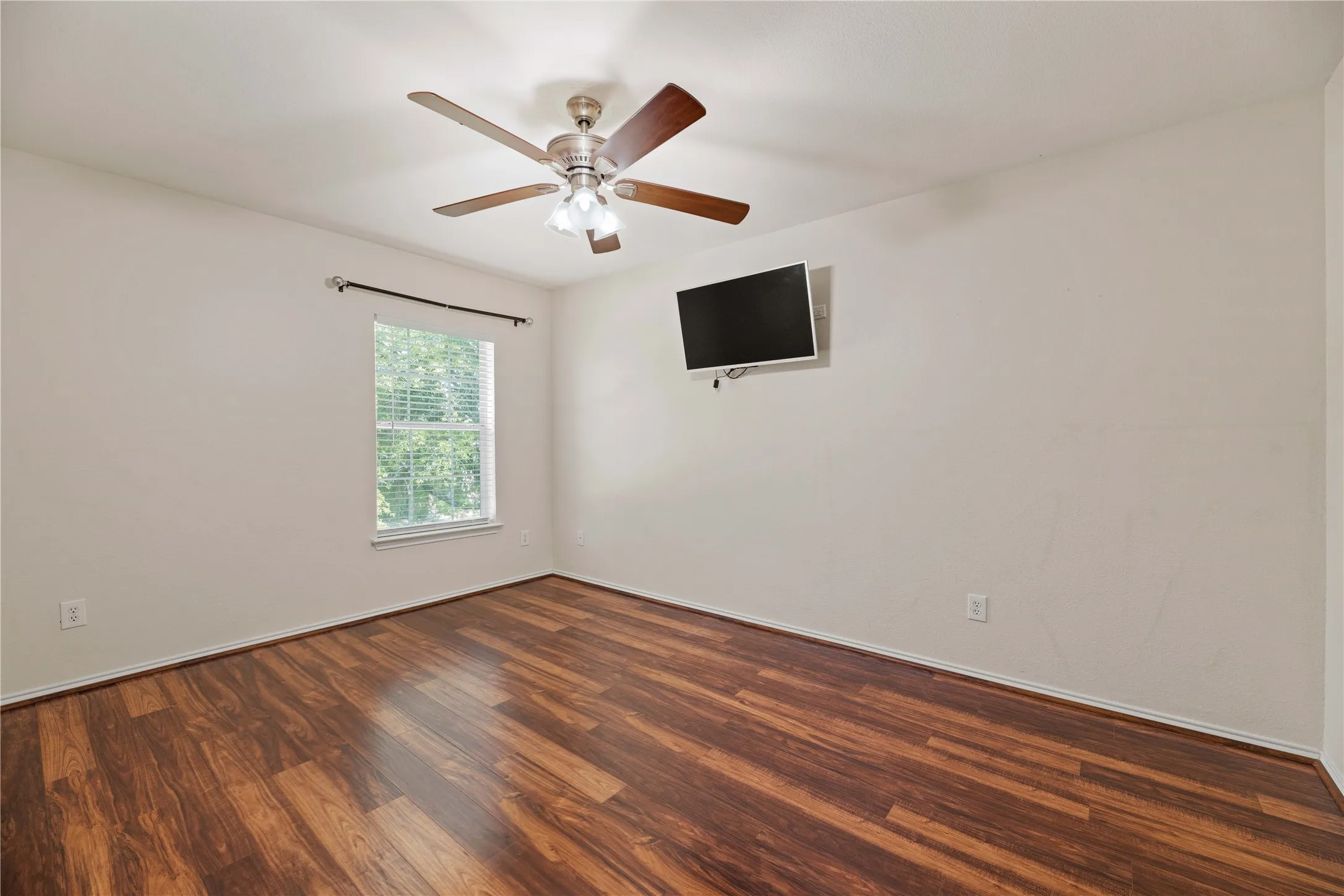 Wood-finish flooring, ceiling fan with integrated lighting, single window with blinds, wall-mounted display