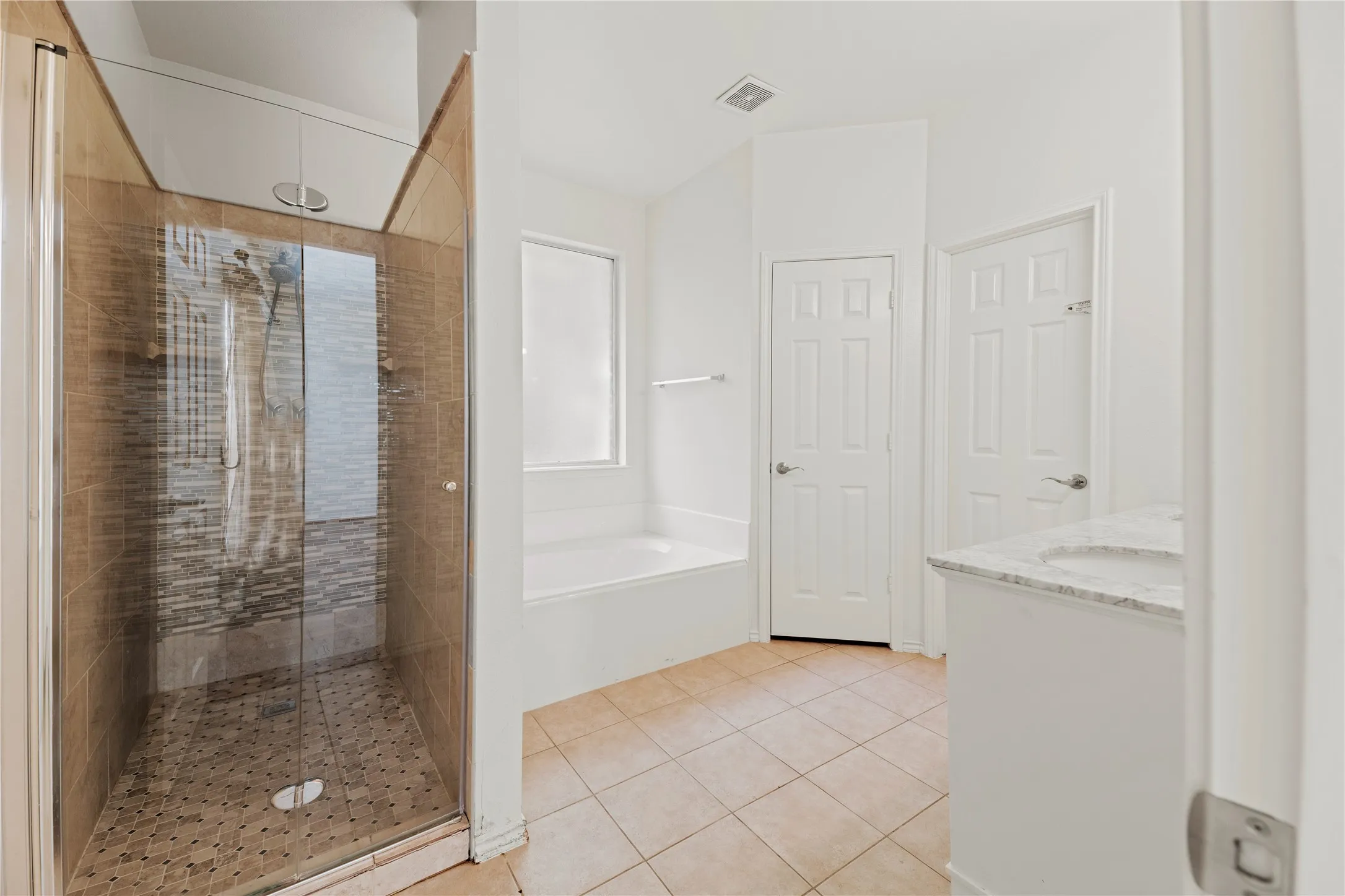 Spa-inspired primary bathroom featuring a walk-in shower with glass enclosure and stone-look tile surround, dual vanities with marble-look countertops, ceramic tile flooring, and abundant natural light from a large window