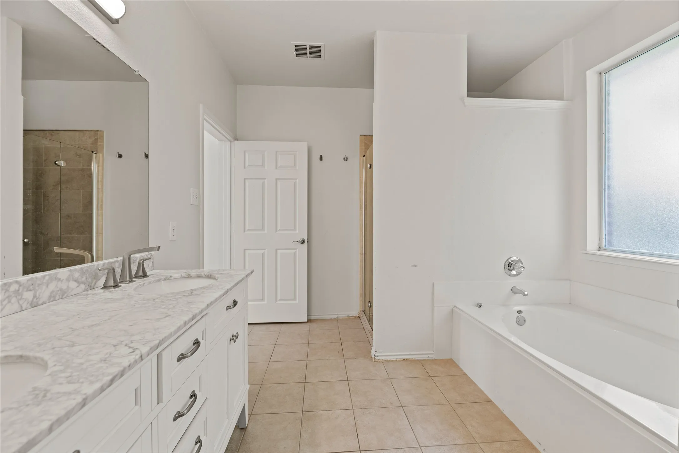 Spa-inspired primary bathroom featuring a dual-sink vanity with marble-finish countertop, white cabinetry with brushed nickel hardware, a separate soaking tub, walk-in shower with glass enclosure and tile surround, and abundant natural light from a large window