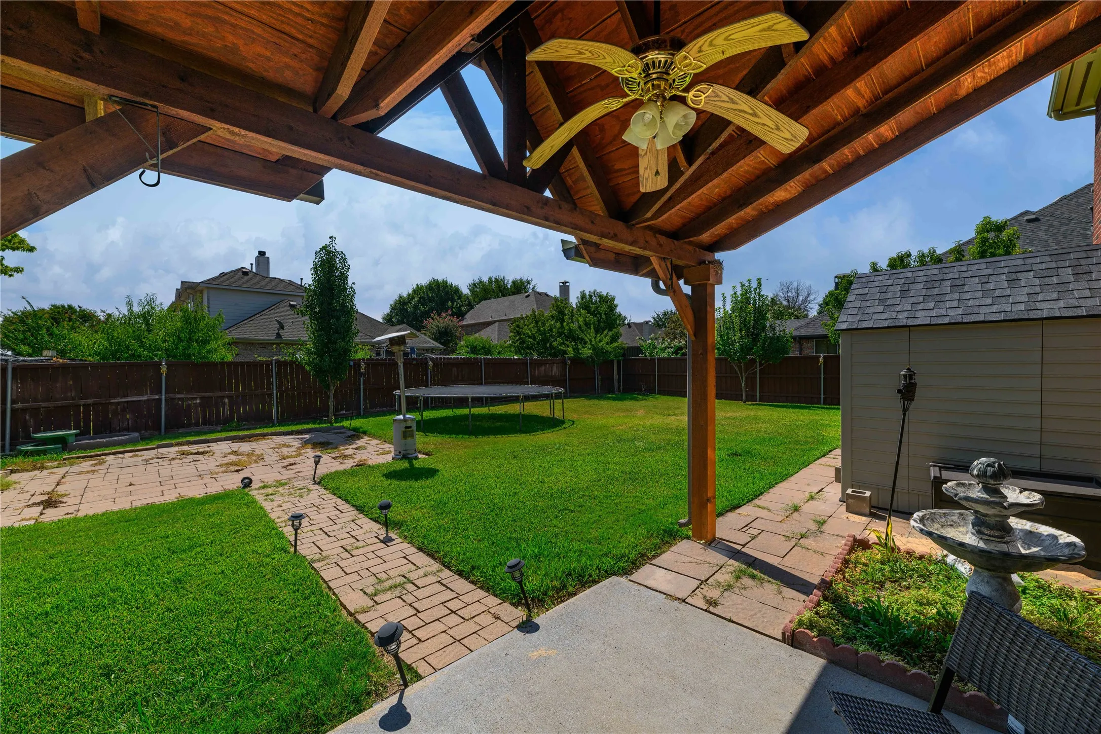 Expansive backyard featuring a covered patio with exposed wood beams and a ceiling fan, a storage shed, and a stone-paver​​‌​​​​‌​​‌‌​‌‌​​​‌‌​‌​‌​‌​​​‌​​ walkway