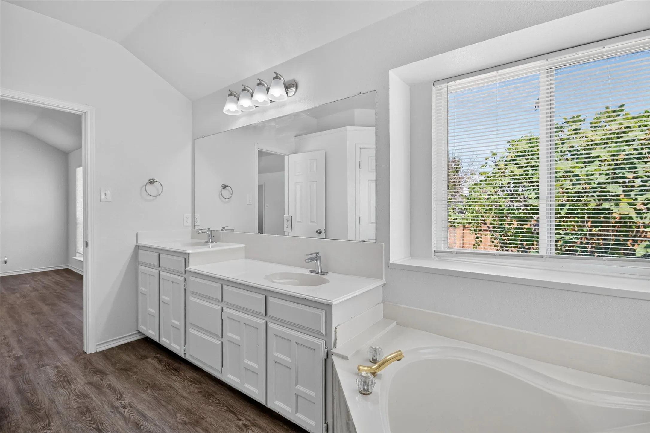Dual vanity with undermount sinks and chrome fixtures, large integrated mirror, recessed soaking tub with gold-tone fixtures, and window with horizontal blinds