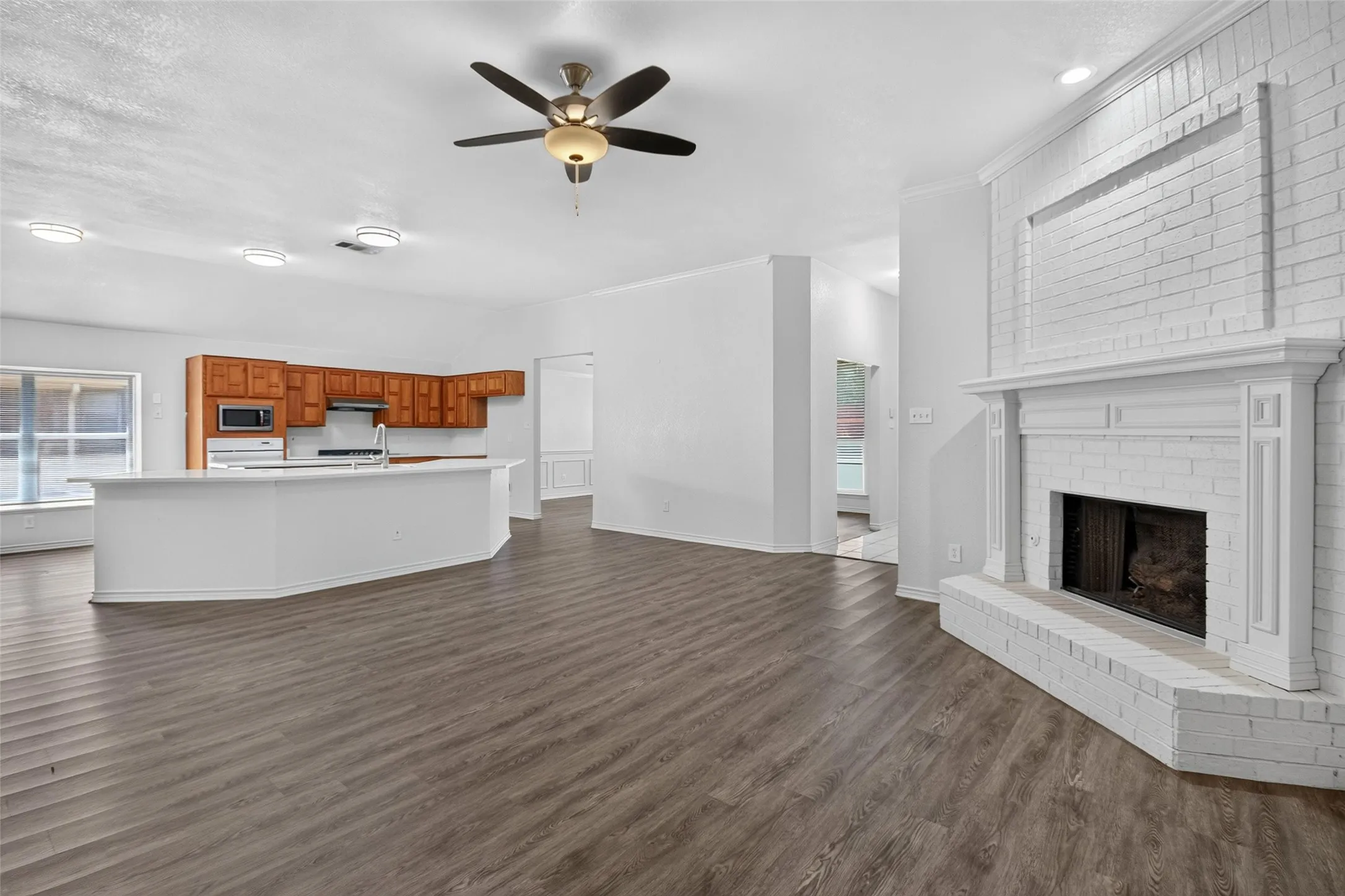 Spacious living area featuring wood-finish flooring, a white-painted brick fireplace with a mantle, and an open-concept kitchen with wood cabinetry and a central island