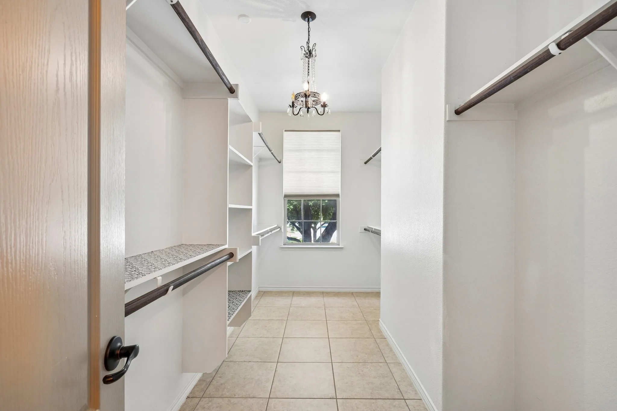 Spacious walk-in closet featuring built-in shelving, hanging rods, tile flooring, a window with a roman shade, and a decorative chandelier