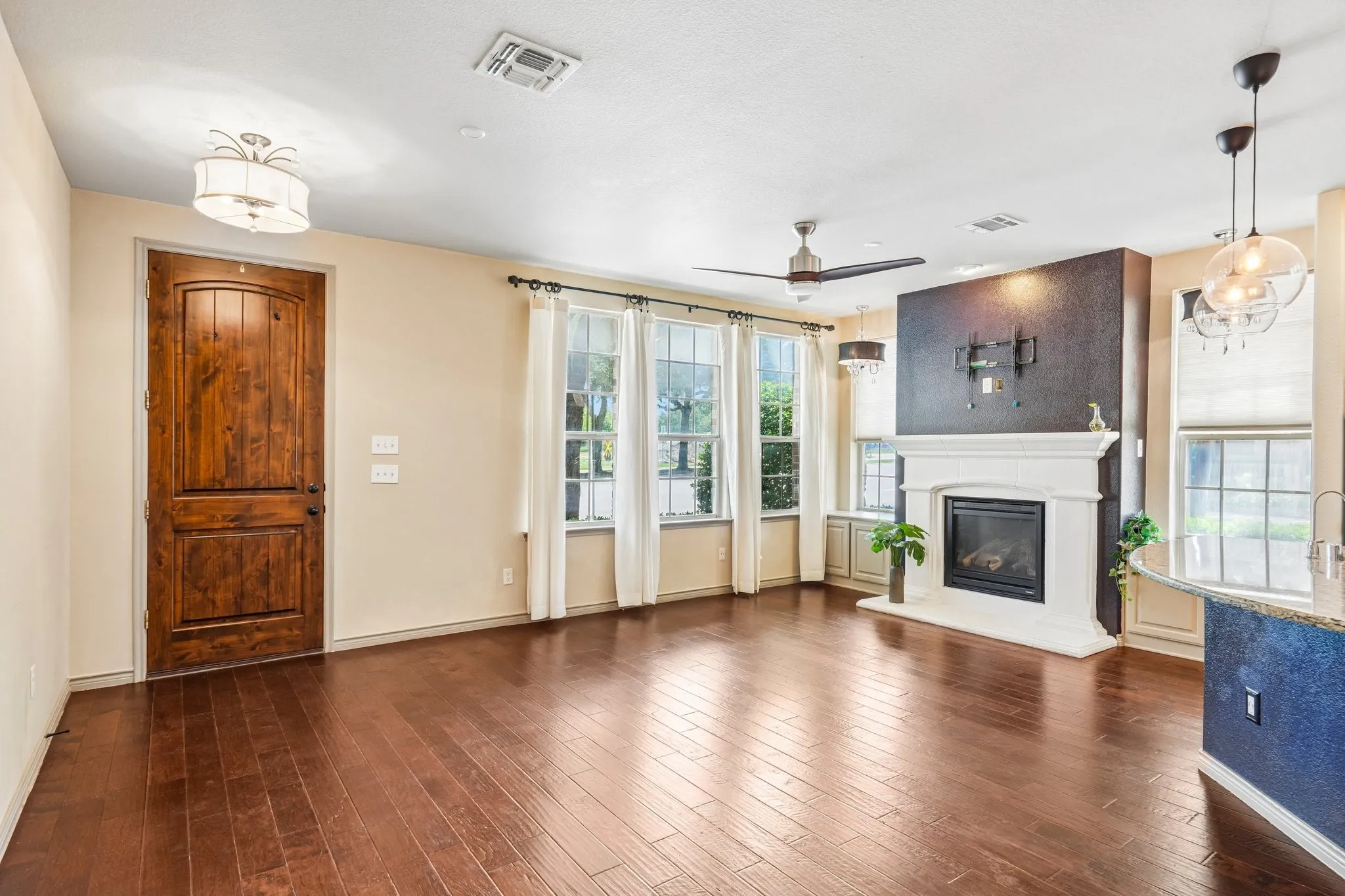 Rich wood-finish flooring, a prominent fireplace with a white mantel, multiple windows with white trim, a decorative entry door, and a built-in ceiling fan with lighting