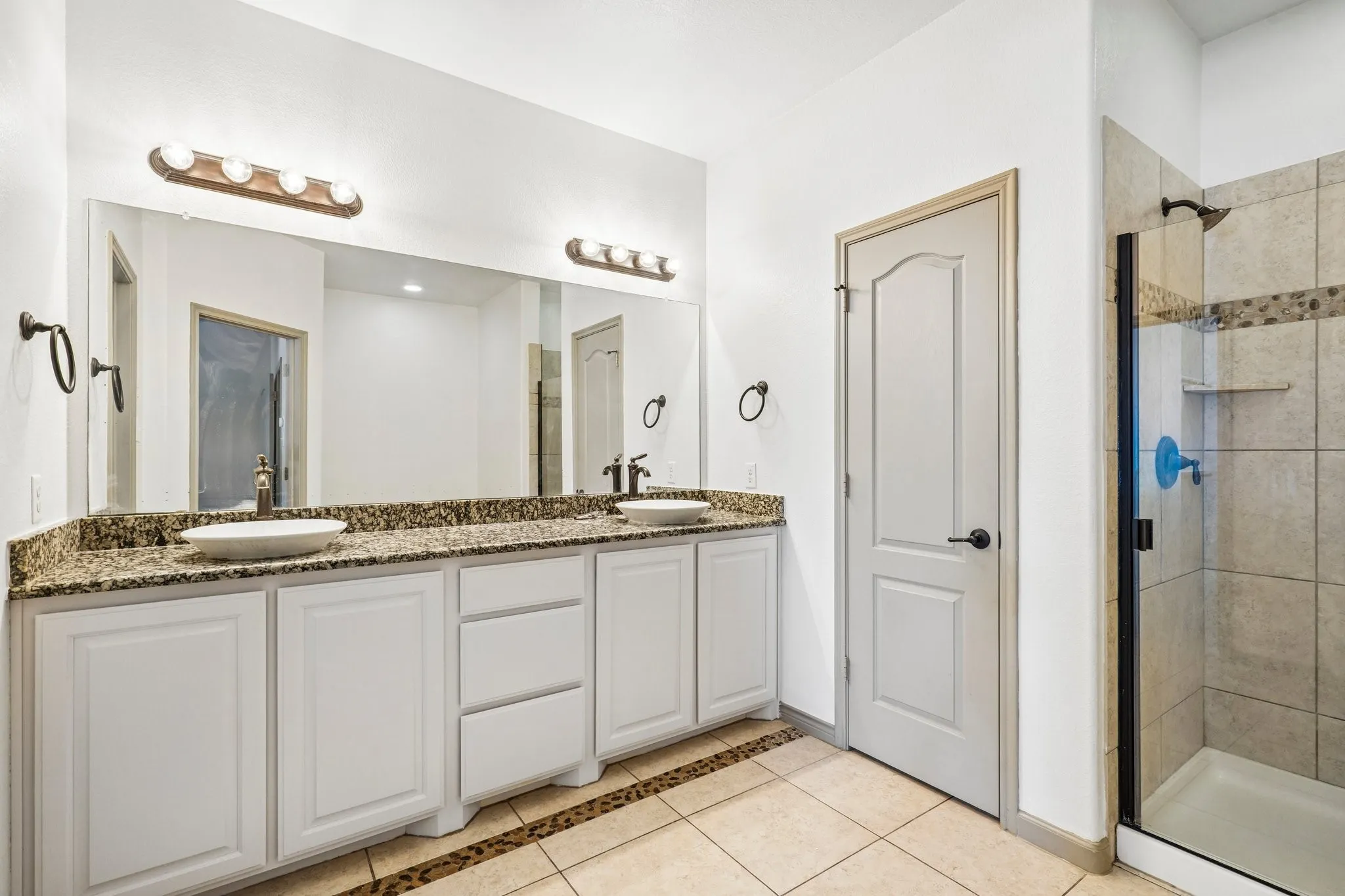 Dual vessel sinks, granite-finish countertop, white cabinetry with bronze hardware, tiled shower with glass enclosure, and neutral tone tile flooring