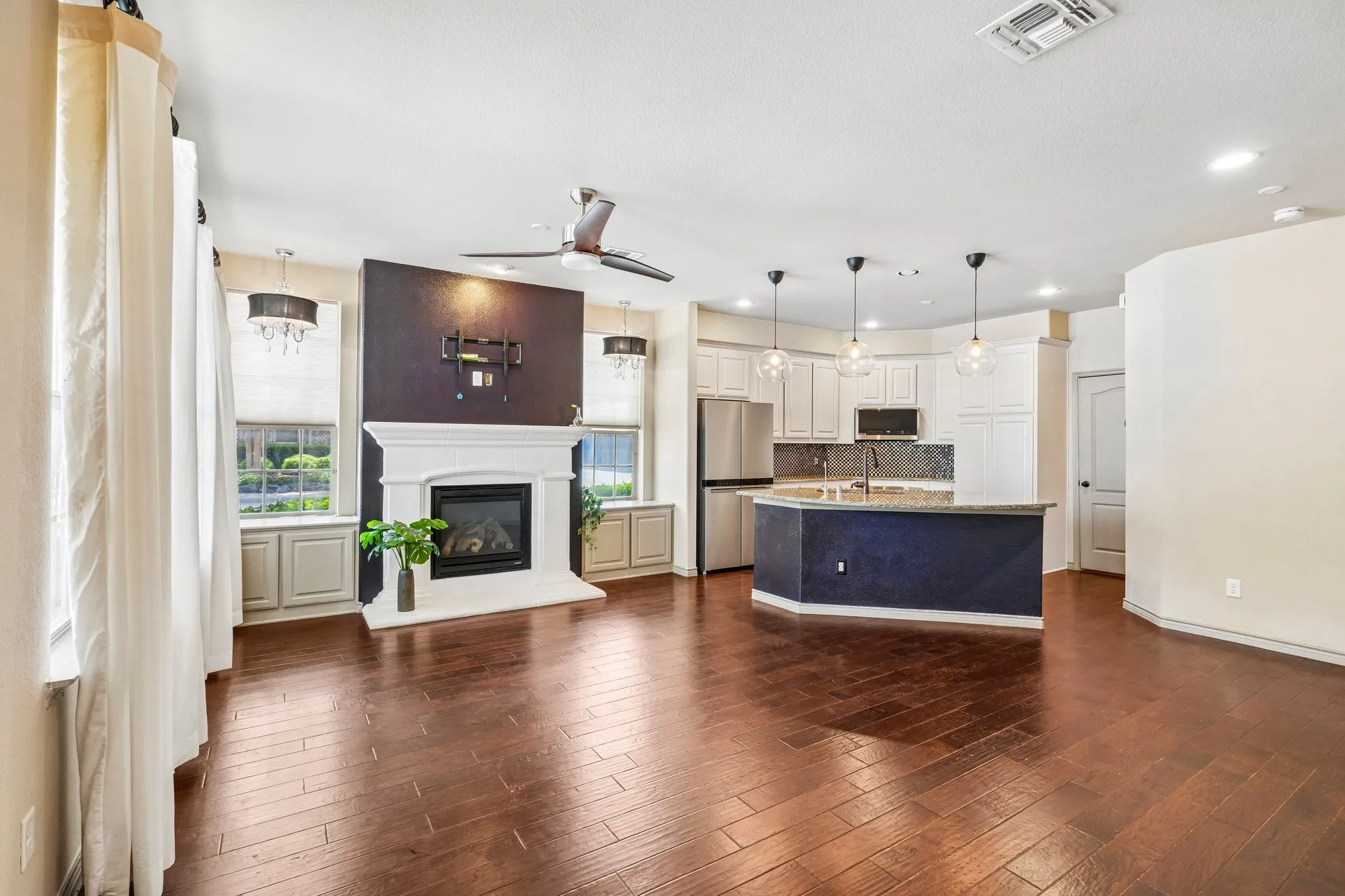 Expansive living area featuring rich wood-finish flooring, a white mantel fireplace, and a ceiling fan