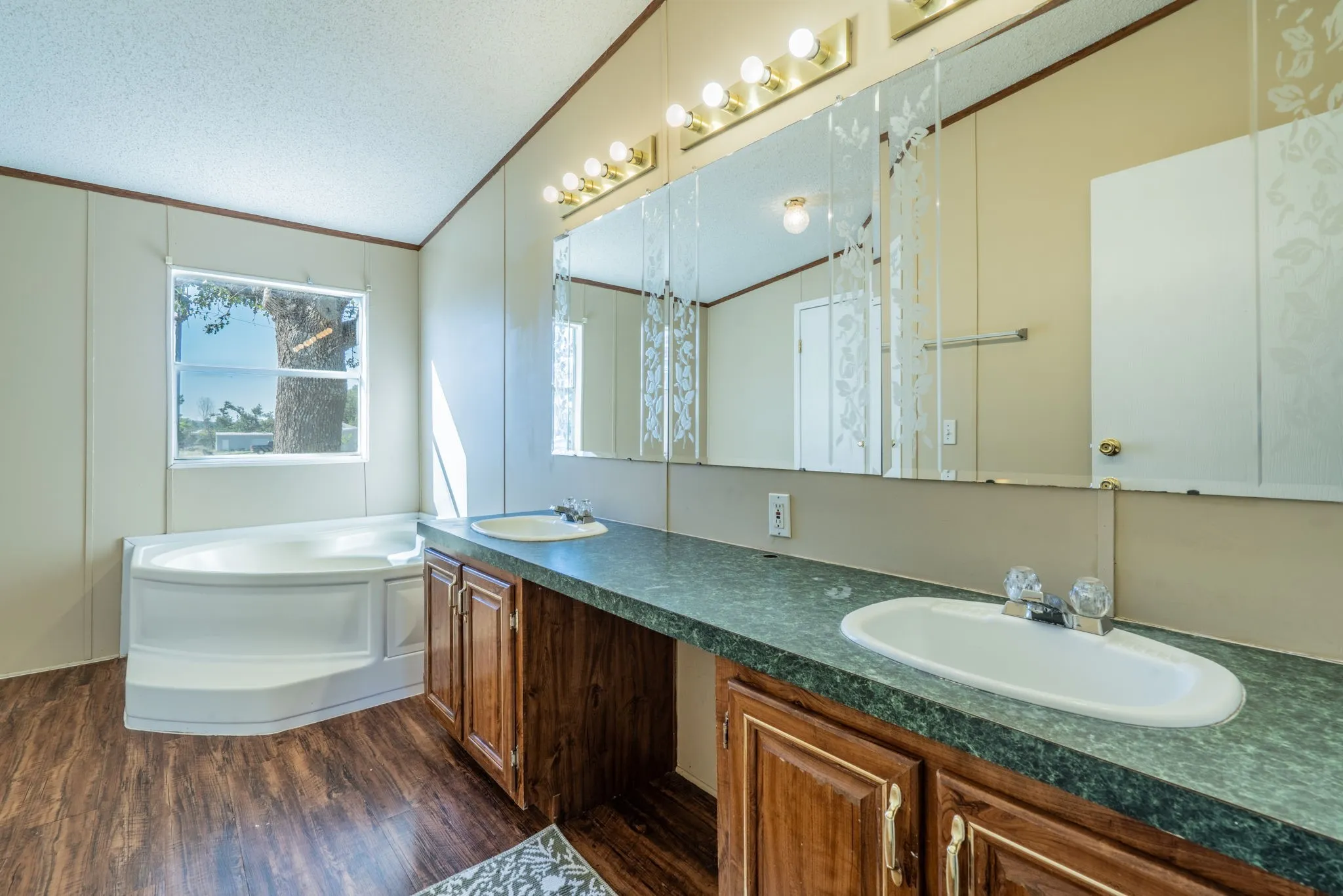 Primary Bathroom: Spacious bathroom featuring a dual-sink vanity, wood cabinetry, decorative mirrors, and a corner soaking tub beneath a window.