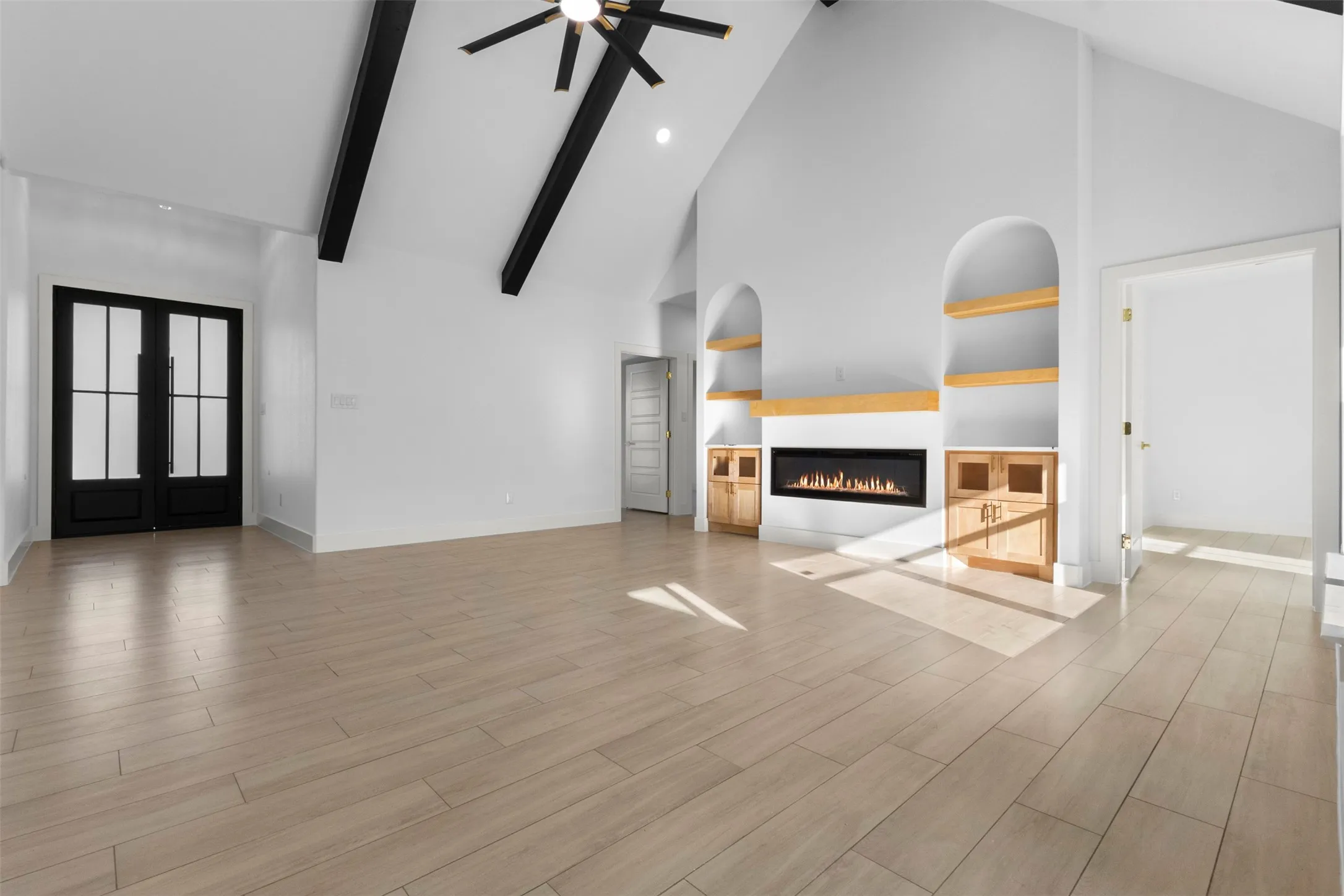 Vaulted ceiling with exposed beams, light wood-finish flooring, built-in fireplace, custom arched shelving, and black-framed double entry doors