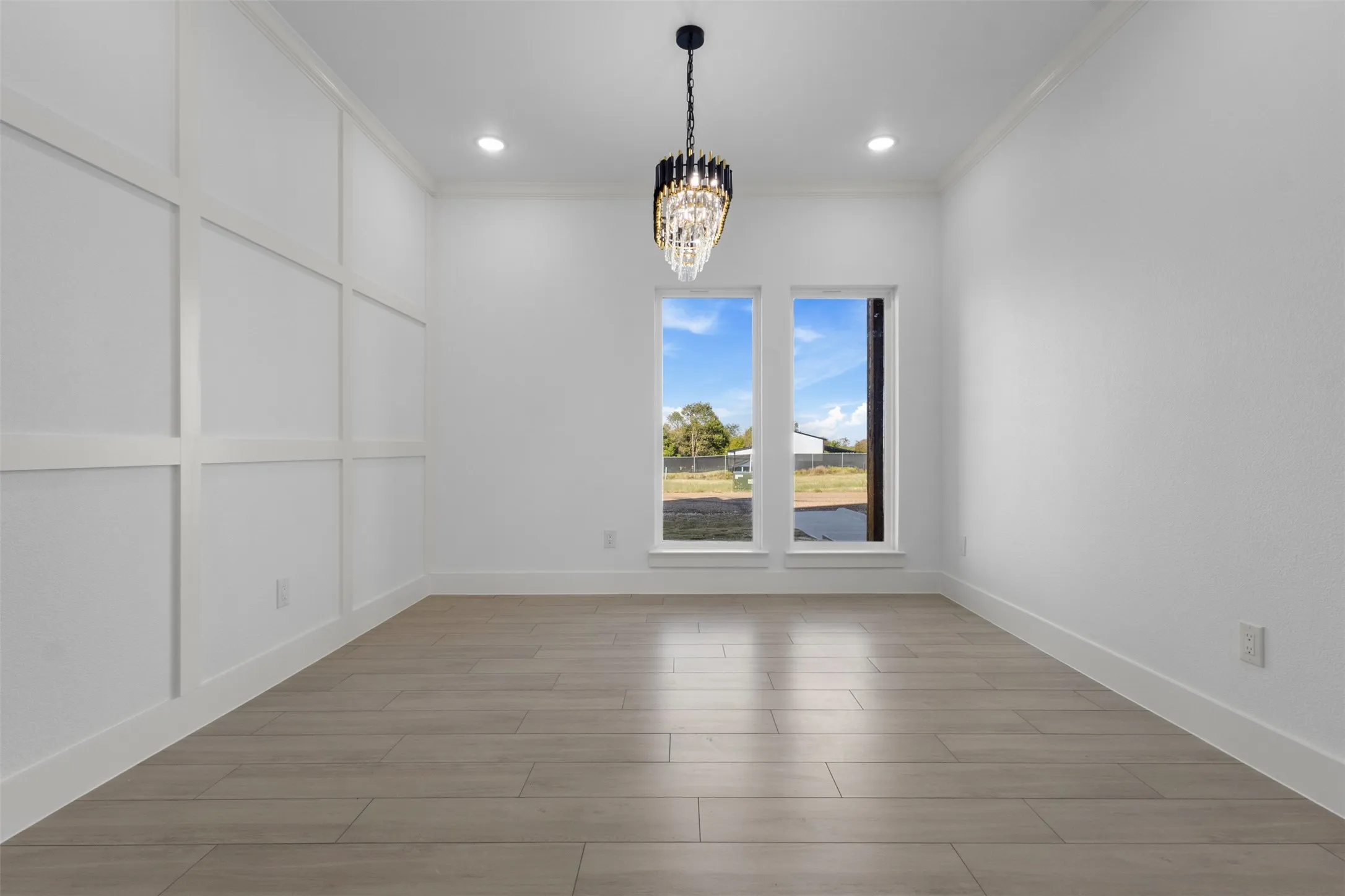 Recessed lighting, elegant chandelier, wood-finish flooring, two large windows, and a decorative paneled accent wall
